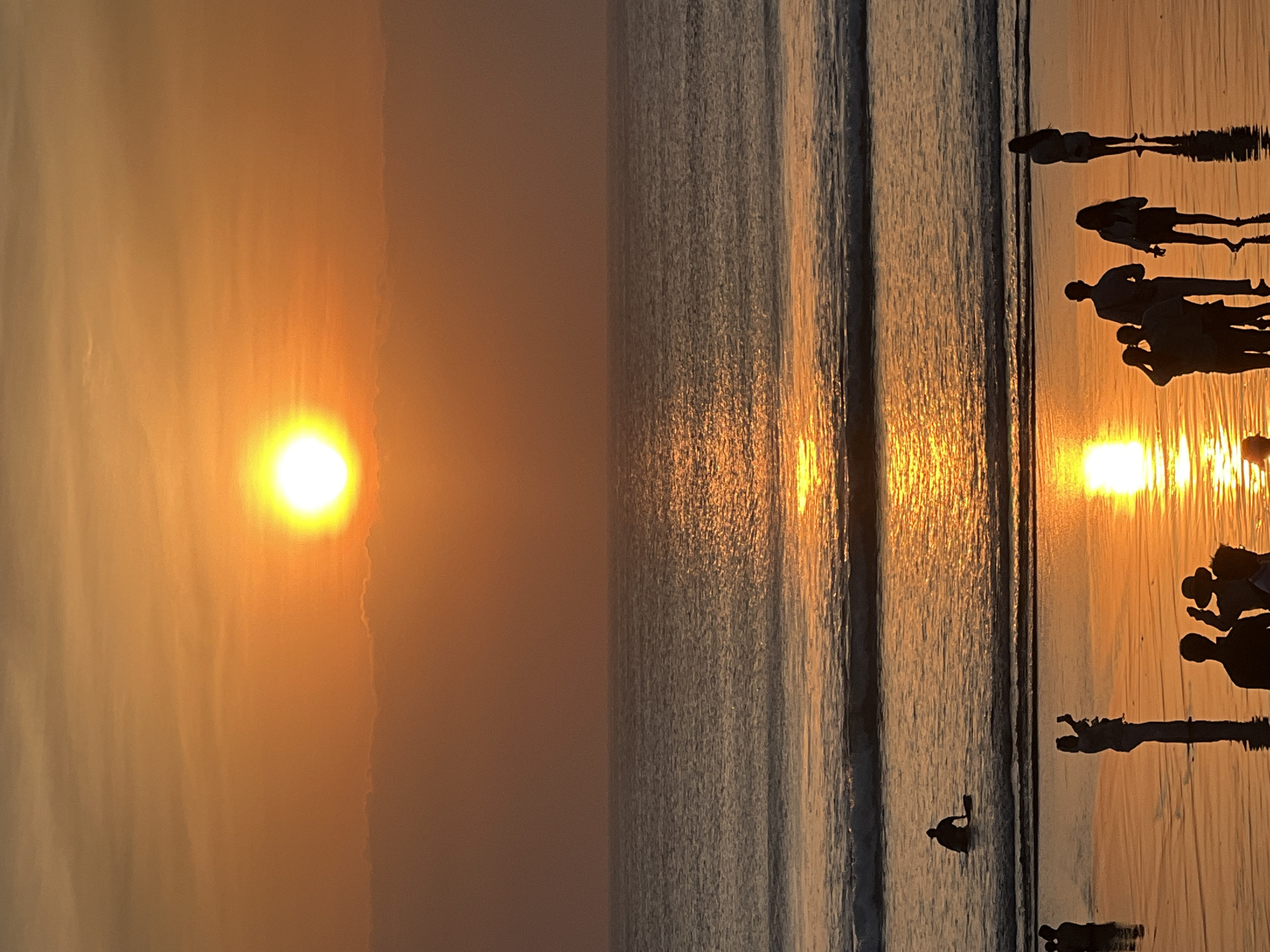 A sunset scene at Kuta Beach, Indonesia. The sun, a bright orb, is low in the sky, casting a warm orange glow over the water and beach. Silhouettes of people are visible, some standing on the wet sand, others in the water. One person is sitting on a surfboard in the ocean. The waves are gentle, reflecting the sunlight. The scene is peaceful and serene, with a sense of calm as the day comes to an end.