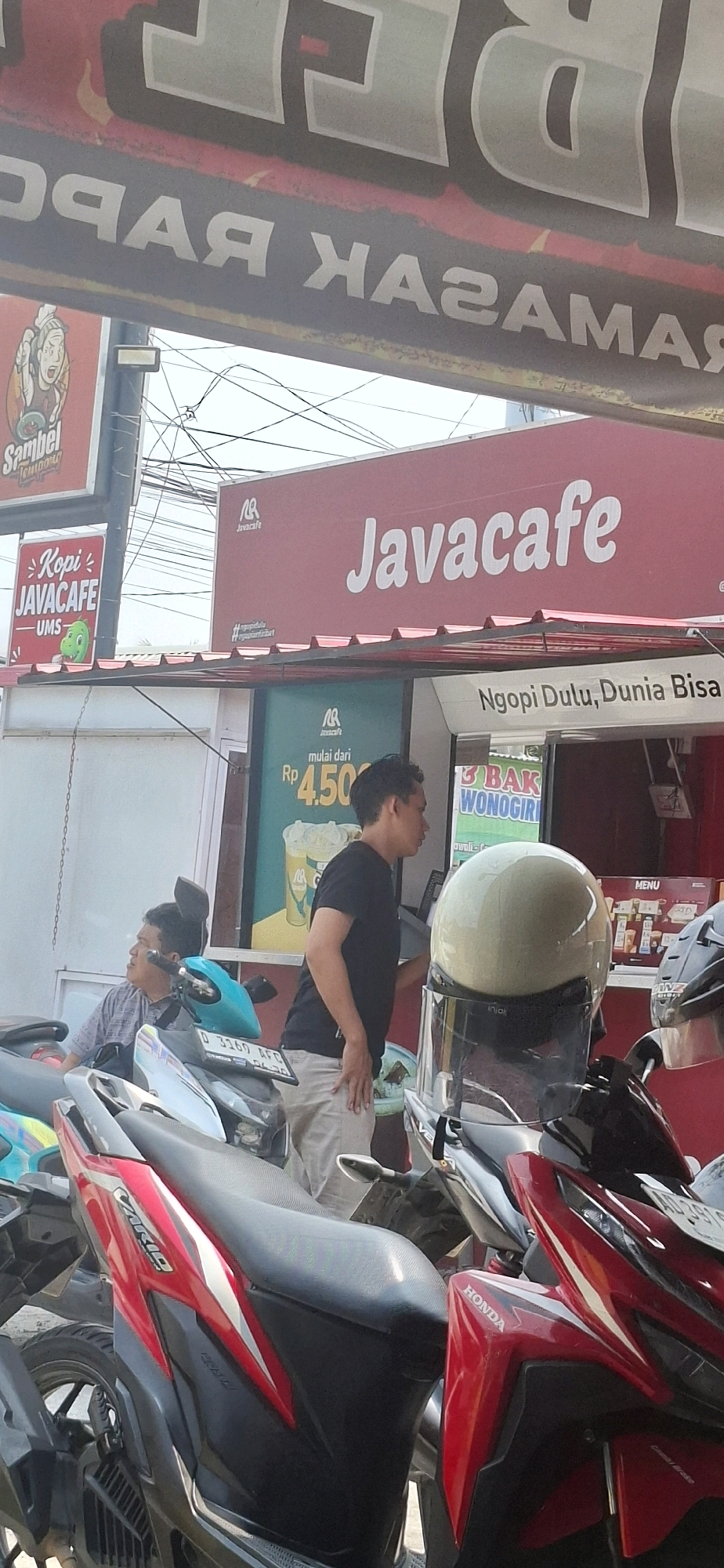 In Pabelan, Indonesia, a busy street scene unfolds outside a "Javacafe" establishment. The cafe features a prominent red awning with the "Javacafe" logo and text in white. Below the awning, a green sign advertises "mulai dari Rp 4.500," suggesting a special offer. To the right, a white sign reads "Ngopi Dulu, Dunia Bisa," a slogan likely encouraging patrons to relax and enjoy coffee.
Two men are present. One, wearing a grey striped shirt, sits on a teal-colored motorcycle. The other, in a black t-shirt and light-colored pants, stands near a white helmet placed on a motorcycle. Several motorcycles are parked in the foreground, including a red and black Honda with visible "HONDA" branding. The environment appears to be an outdoor seating or ordering area, with power lines visible overhead. Other signs and banners are present but not entirely legible, including one with a cartoon character and the word "Sambel."