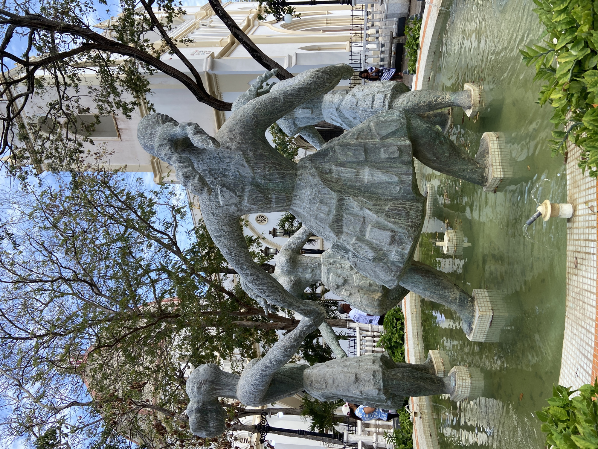 The image depicts a bronze sculpture of children playing in a fountain in a park. The statue is made up of four figures holding hands and moving in a circle. The fountain is surrounded by greenery. In the background, there is a white building with a bell tower. The sky is blue with some clouds. The scene is located in Porlamar, Venezuela.