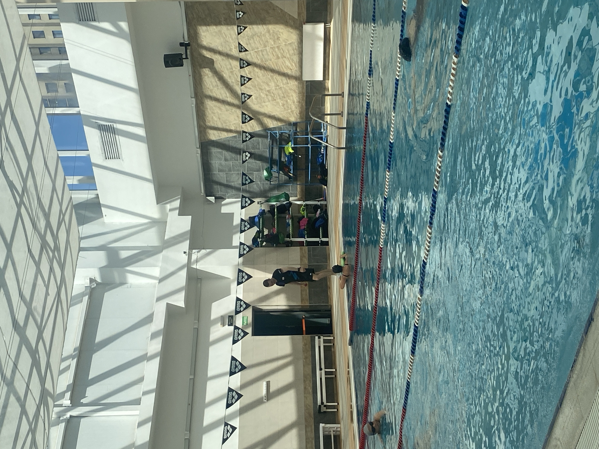 A high-angle photograph shows an indoor swimming pool at the Water Sports Palace in Yekaterinburg, Russia. The pool has lane markers of white, red, and blue. Three people are in the water, swimming. A man stands at the edge of the pool, near an open doorway. The surrounding area includes flags with the word "Arena" printed on them, lockers, benches, and a rack for swim equipment. The architecture features a large window with a geometric pattern.