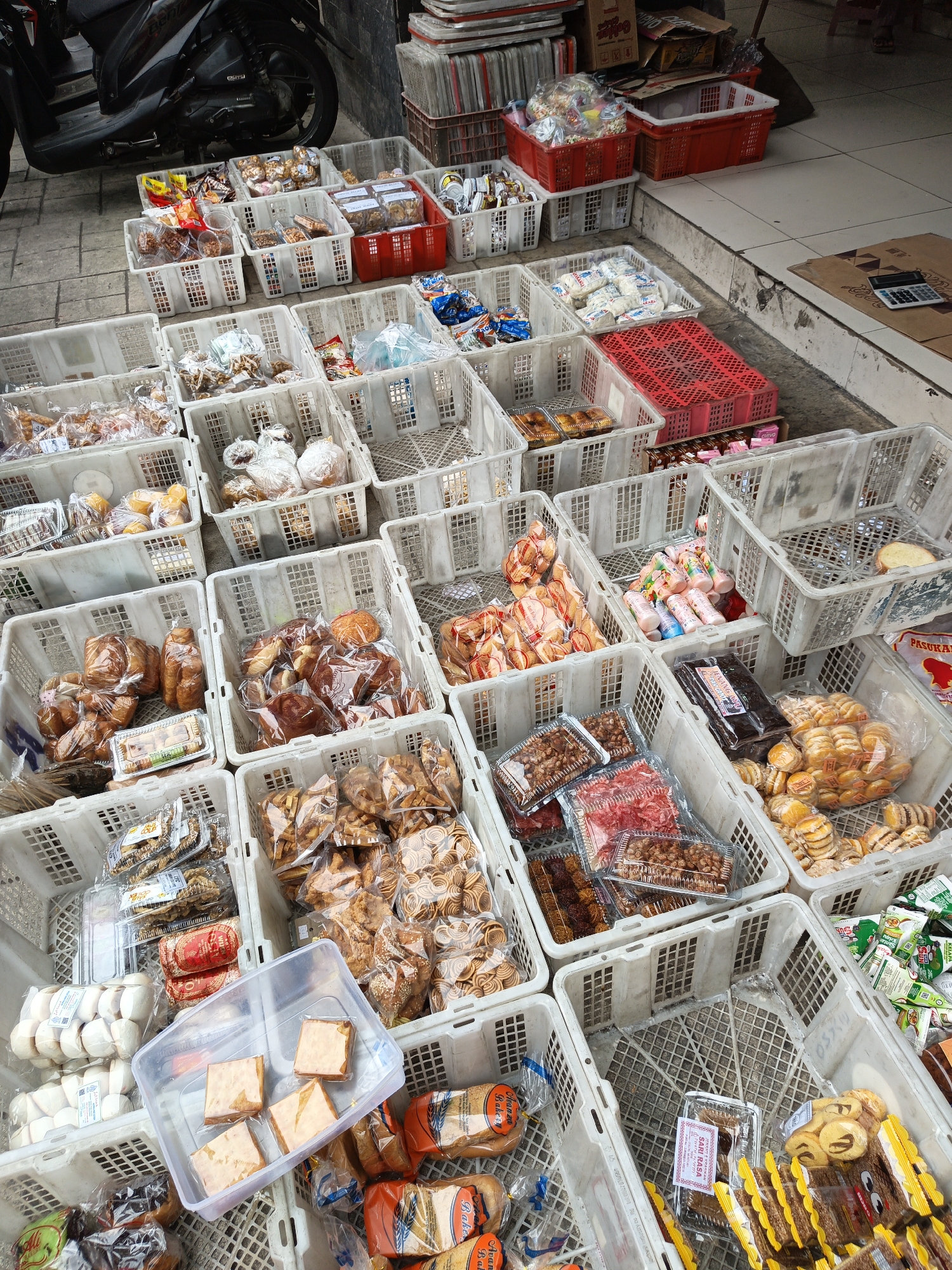 The image depicts an outdoor stall in Klaten, Indonesia, displaying a wide variety of packaged snacks and baked goods. The stall is set up along a sidewalk with a tiled surface, and a motorcycle is partially visible on the left, suggesting a roadside or market setting. The products are organized in white plastic crates of various sizes, with some red crates also present.
The crates are filled with an assortment of items, including:
* Individually wrapped pastries and bread, some in clear plastic bags, others in branded packaging.
* Bags of crispy snacks, such as crackers and fried dough, some with intricate spiral patterns.
* Smaller, individually packaged sweet treats, possibly cookies or candies.
* A clear plastic container holding several square pieces of a light brown food item, possibly cake or a dense pastry.
* Some items are in clear plastic boxes or trays, allowing for visibility of their contents.
* In the background, stacked crates and boxes, along with some loose items, indicate a commercial environment.
The lighting suggests it is daytime, with natural sunlight illuminating the scene. There are no visible people actively engaged in transactions in this specific frame. The overall impression is a street-side vendor with an abundance of affordable packaged snacks.