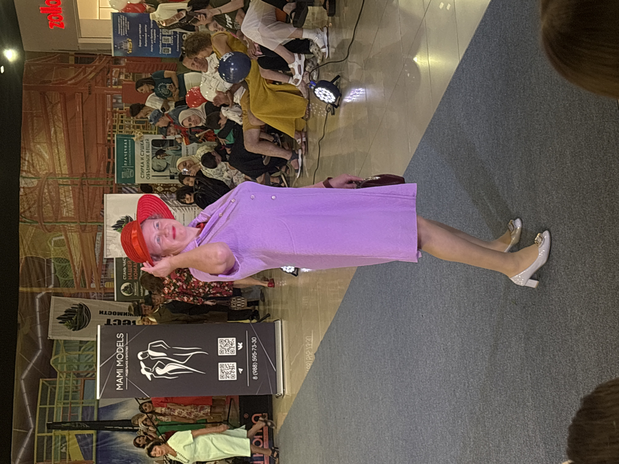 The image depicts a fashion show event taking place indoors, likely in a shopping mall or community center. A woman in a lavender dress and a red hat is walking on a carpeted runway, looking towards the right side of the frame. She is holding a dark purse in her right hand and adjusting her hat with her left hand. In the background, several people are seated in an audience area. Behind the audience, there are banners and displays related to the event, including one for "MAMI MODELS." The environment appears to be well-lit, with some stage lighting visible on the floor. There is no visible indication of the specific location being in Tri Potoka, Russia. The scene is a typical fashion event with models, audience members, and promotional materials.