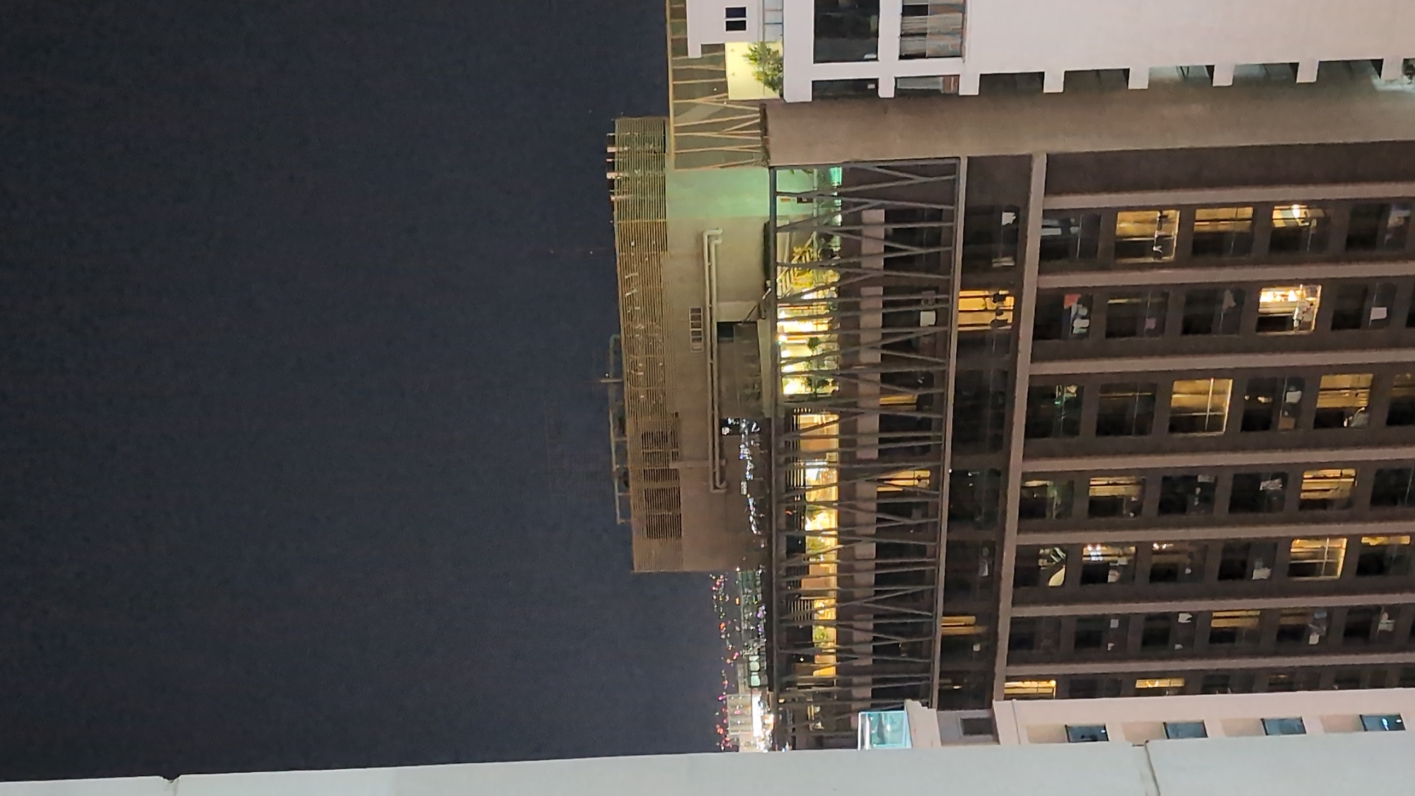The image captures a nighttime view of a modern high-rise building in Nha Trang Ward, Vietnam. The building is illuminated from within, with many windows showing warm, yellow light, suggesting occupancy. Laundry is visible hanging from several balconies, adding a domestic detail to the urban landscape. The building's facade features a geometric pattern of vertical and diagonal elements. In the background, distant city lights are visible against the dark night sky, indicating a developed urban environment. The overall scene depicts a bustling city at night, with residential life evident in the illuminated apartments.