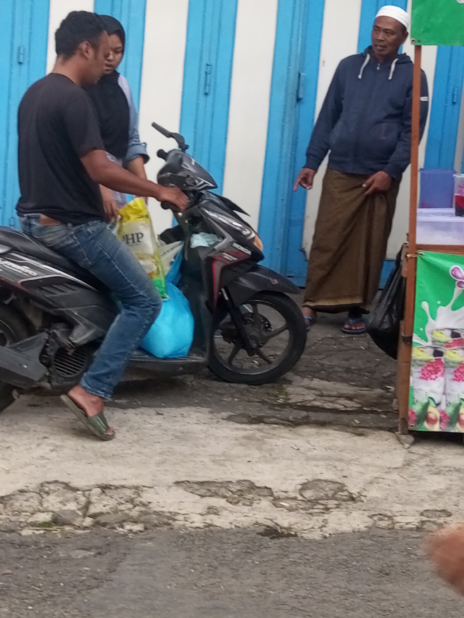 In Pandansari, Indonesia, two men and a woman are near a motorcycle and a vendor's stall. The man in the foreground, wearing a black t-shirt and blue jeans, is seated on a black motorcycle. Next to him, a woman wearing a black hijab and a light-colored shirt stands behind him. Further to the right, another man, wearing a white cap, a dark blue hooded jacket, and a brown sarong, stands near a food stall with blue and white striped doors in the background. The stall displays colorful drinks and fruit. The ground is paved with a rough, textured surface.