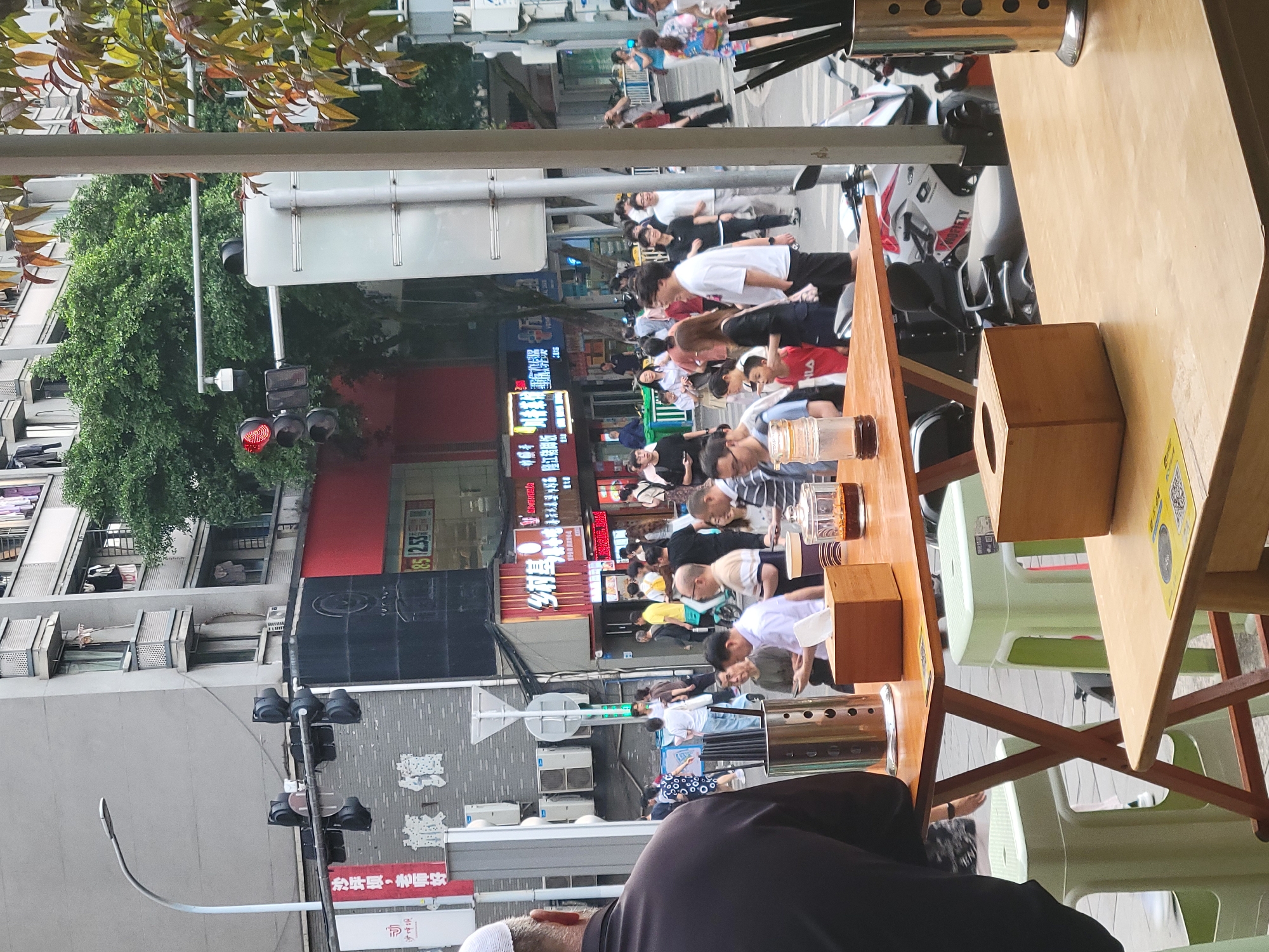 The image shows an outdoor dining area in the Shapingba District of Chongqing, China. A wooden table and green plastic chairs are in the foreground. People are seated and appear to be eating. In the background is a busy street scene with people crossing at a crosswalk. There are shops and businesses with Chinese signage visible. Traffic lights are red, suggesting cars must stop for the pedestrians. There are parked motorcycles along the curb. A lamppost is on the left, and trees are partially visible at the top and left of the frame.
