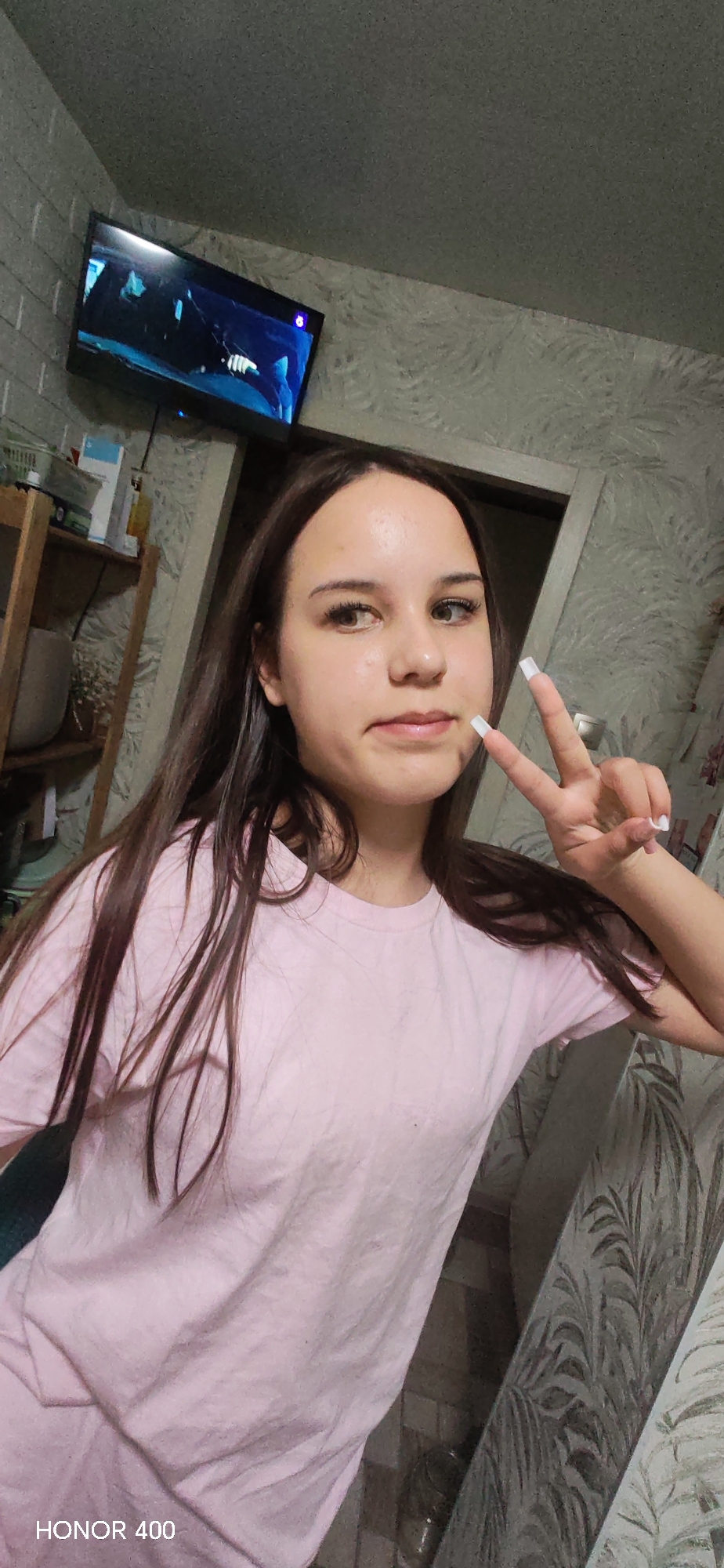 A young female, likely in her late teens or early twenties, is featured in a selfie taken indoors, presumably in Noviy, Russia. She has long, straight dark brown hair that falls over her shoulders, fair skin, and light-colored eyes. Her expression is neutral with a slight hint of a smile, and she is making a peace sign with her right hand, showcasing long, square-tipped white nails. She wears a plain, light pink short-sleeved t-shirt.
The setting appears to be a casual home environment, possibly a living room or bedroom. Behind her, a flat-screen television is mounted on a wall with grey brick-patterned wallpaper or tiles, displaying what looks like a dark scene with blue lighting, possibly from a movie or TV show. To the right of the person, the wall is covered with a light-colored wallpaper featuring a subtle leaf or fern pattern. A wooden shelving unit is visible on the left side of the frame, holding various household items, including bottles and a grey pot or vase. A door frame is also partially visible behind her right shoulder.
The lighting suggests it is evening or night, relying on indoor artificial light sources. The overall activity is a posed snapshot, capturing a relaxed moment. A visible watermark in the bottom left corner of the image reads "HONOR 400," indicating the device used to capture the photograph.