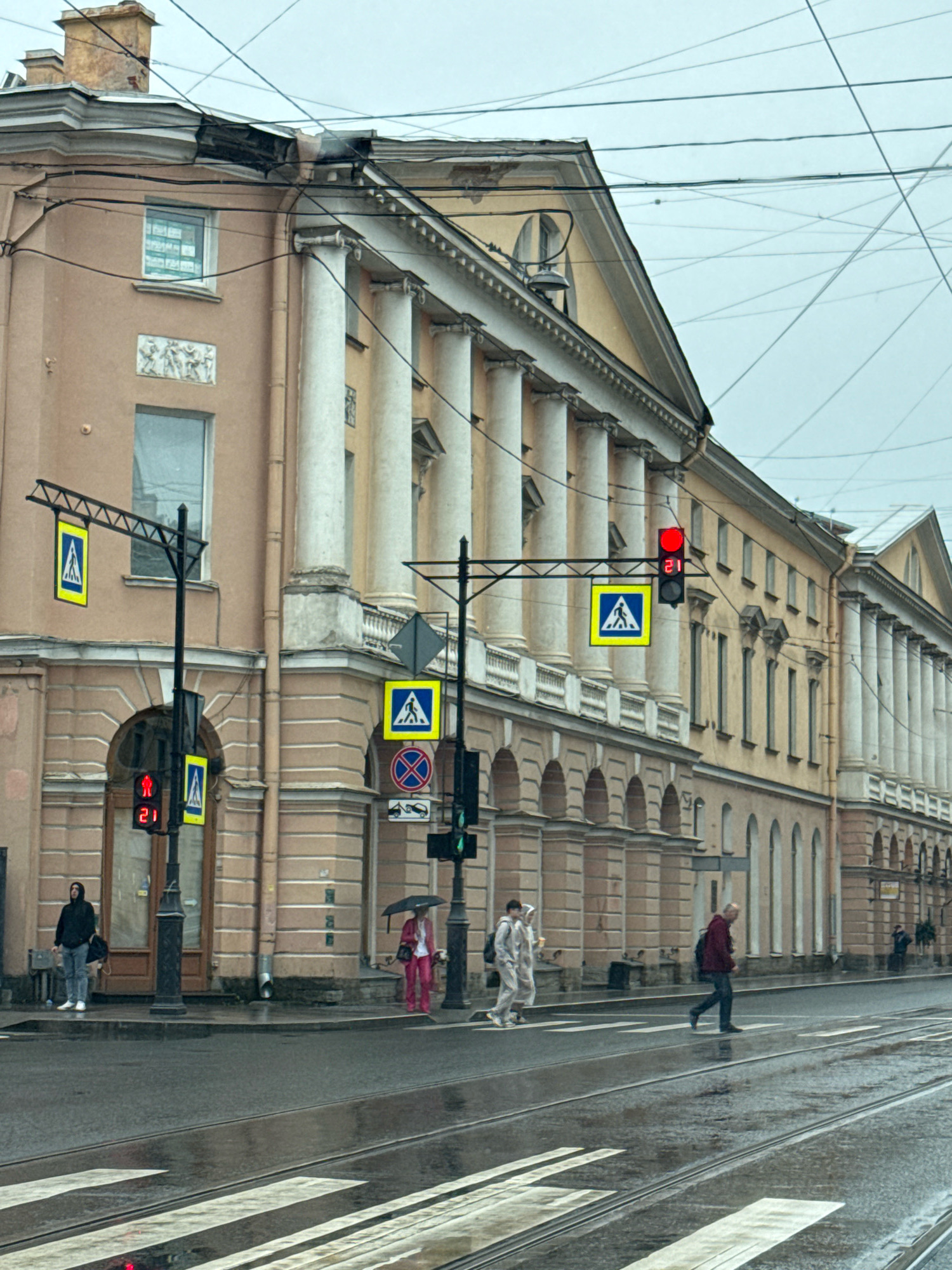 The image depicts a street scene in Saint Petersburg, Russia, on a cloudy, rainy day. The street is wet, with reflections visible on the asphalt, and tram tracks are present. In the foreground, a pedestrian crossing is marked by white stripes.
On the right side of the street, a man in a dark red jacket and dark pants is walking across the pedestrian crossing. To the left of him, two figures in light-colored clothing are walking together. Further left, a person in a dark hoodie and jeans stands on the sidewalk, holding a bag. Across the street, a person in a pink suit and a dark umbrella is standing on the sidewalk.
Traffic lights and pedestrian crossing signs are visible. A red pedestrian signal with a countdown timer showing "21" is illuminated. Several buildings with classical architecture, featuring columns and arched windows, line the street. Overhead, numerous electrical wires crisscross the sky. A damaged section of a building's roof and gutter is visible on the upper left.