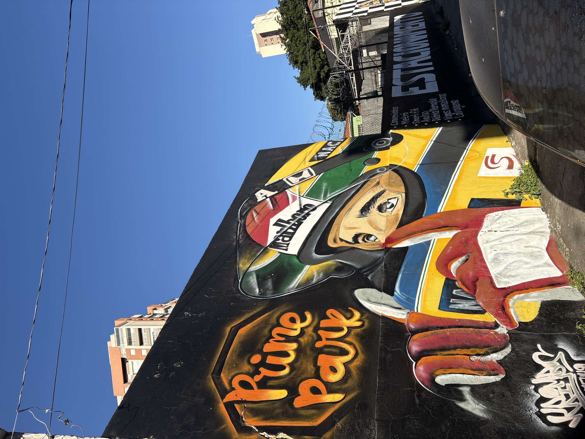 This is a street-level, medium-shot photograph of a vibrant mural painted on a black wall in São Paulo, Brazil. The mural features a stylized portrait of a race car driver wearing a helmet with a "Marlboro" logo. The driver's face is depicted with large, expressive eyes, and their gloved hands are prominently displayed in the foreground, one pointing forward. To the left of the driver, the words "Prime Park" are spray-painted in large, orange, stylized lettering with a yellow outline. The mural incorporates bold colors like yellow, red, green, and blue, characteristic of street art.
Behind the mural, the urban environment is visible. A clear blue sky dominates the upper portion of the image. In the background, there are buildings, including a modern, orange-tinted apartment building and a lighter-colored building further in the distance. A chain-link fence with barbed wire runs along the top of the wall, separating the mural from the area behind it, which appears to be a parking lot or an area with buildings. Black and white checkered patterns are visible on a wall to the right, suggesting a racing or automotive theme continuing beyond the main mural. Text in white spray paint on the black wall to the right of the driver's helmet reads "ESTACIONAMENTO" and a bulleted list including "Conveniados," "Metro," "Smart fit," "Aviso," "Diária/Permante," "Vagas mensais," and "Lavagens." A dark-colored car is parked in the foreground, partially obscuring the lower right of the mural and the text. Small green plants are growing at the base of the wall where the mural ends. The overall impression is one of urban decay contrasted with bright, energetic street art.