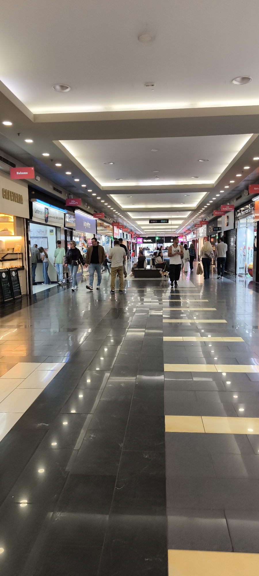 This media file depicts a bustling interior view of a modern shopping mall corridor in Barberà del Vallès, Spain. The scene shows numerous people of various ages walking, standing, and a few sitting on benches along the wide passageway. Shoppers appear to be moving in both directions, either browsing the storefronts or navigating through the mall. A small dog on a leash is visible near a seated group.
The mall features a contemporary design with a bright, clean aesthetic. The ceiling is white, adorned with numerous recessed circular lights and continuous strip lighting along its edges, providing ample illumination. The floor is a striking feature, composed of large, glossy dark grey or black tiles, interspersed with sections of white and bright yellow tiles that create a linear pattern. The highly reflective surface mirrors the overhead lights and the movement of people.
Storefronts line both sides of the corridor. Visible text on the left includes "CAFETERIA," a red sign for "Boheme," and "Loterias y Apuestas del Estado" with the number "74778" on a sign inside. Further down, a store sign reads "Juguetes" (Toys). On the right side, signs for "PS," "Natura," "Amargura Store," and "S.M.A.K." can be observed. A digital clock or display on one storefront shows "11:41," indicating the time. One person in the foreground is wearing a t-shirt with "Vans" printed on it. The overall atmosphere is lively and typical of a commercial center during its operating hours, suggesting late morning or midday.