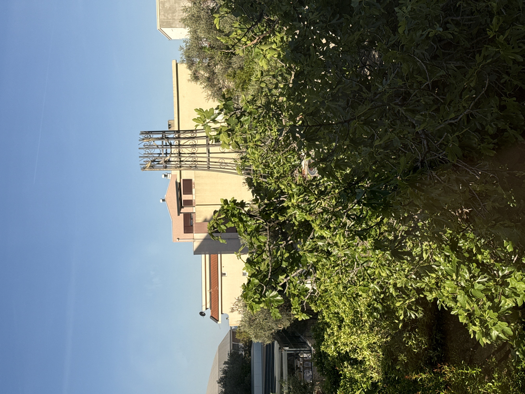 The image shows a residential area, likely in Bar, Montenegro, based on the architectural style and vegetation. A multi-story building with light-colored facades and reddish-brown tiled roofs is visible. An ornate, wrought-iron structure stands against the building. Lush greenery, including trees and bushes, fills much of the right side of the frame. A glass-enclosed structure, possibly a conservatory or greenhouse, is visible at the bottom. The sky is clear and blue.