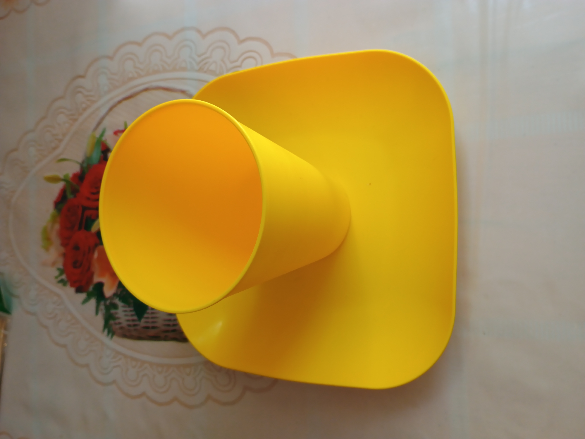 A bright yellow plastic cup sits on a matching yellow square saucer. The saucer has rounded corners and is placed on a table covered with a light-colored tablecloth. Behind the saucer and cup, there is a decorative element, possibly a doily or placemat, with a floral pattern featuring red roses and green foliage. The overall setting appears to be indoors, likely a domestic environment. No specific location in Zaporizhzhia, Ukraine can be determined from the image.