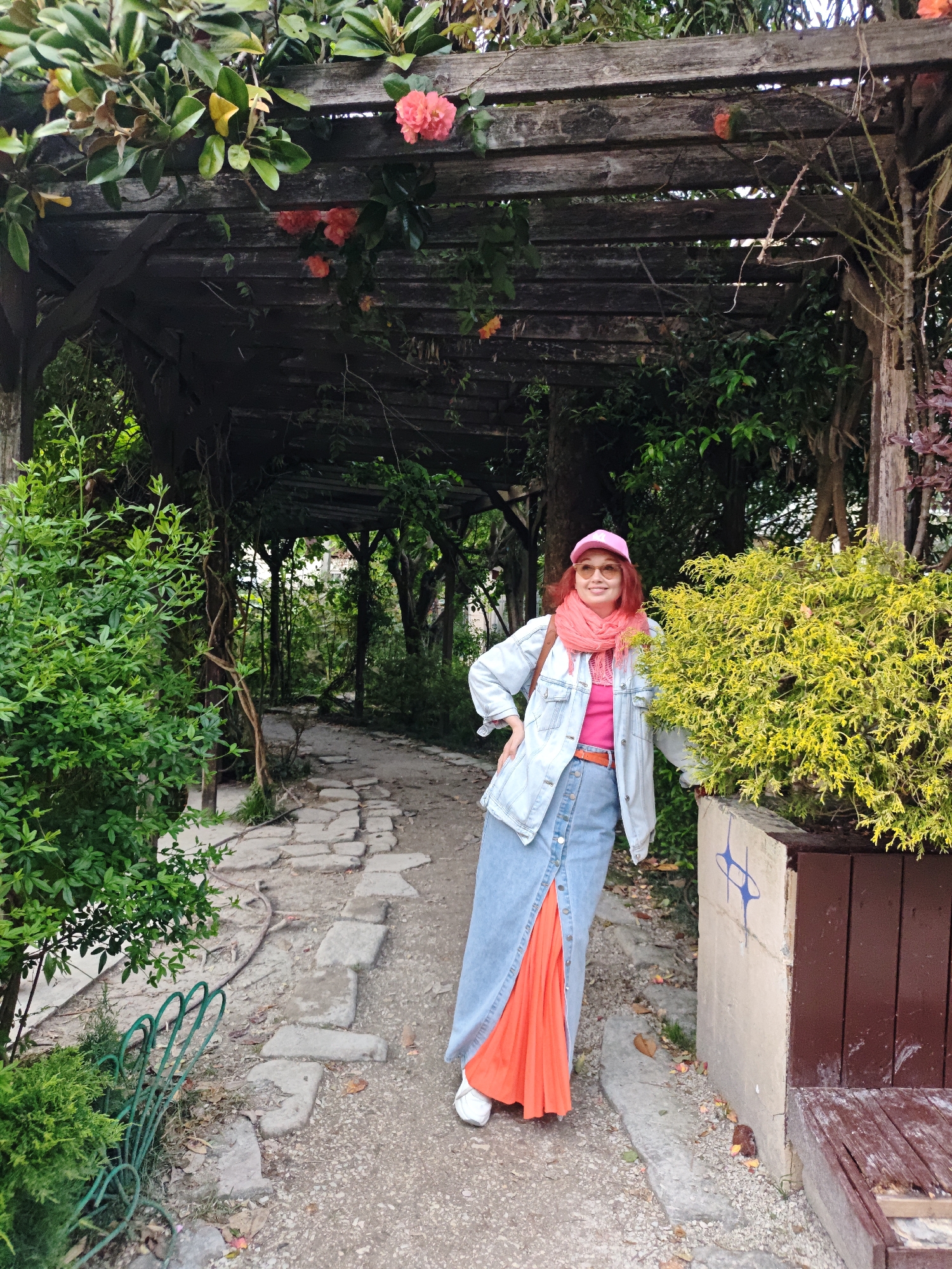 The image depicts a woman standing in a park-like setting. She is fair-skinned, has red hair and is wearing a pink cap, pink scarf, pink shirt, light blue denim jacket, a long denim skirt with orange fabric showing through a slit, and white sneakers. She is leaning against a concrete and wooden planter box with yellow and green foliage, with one hand on her hip and the other on the planter.
Behind her is a stone pathway running through lush greenery and under a wooden pergola covered in foliage and red flowers. The setting appears to be a garden or park.
The location is the city of Sochi, Russia.