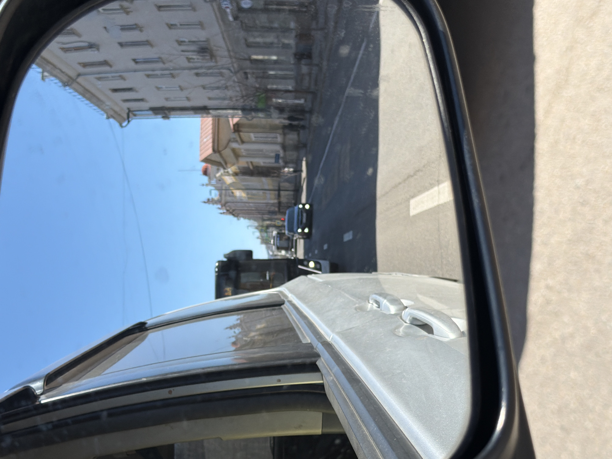 The image captures a street scene as viewed through a car's side mirror, rotated 90 degrees clockwise. The perspective is from inside a silver vehicle, with parts of its door, handle, and roofline visible around the dark frame of the mirror.
Reflected in the side mirror is an urban street under a clear, bright blue sky. The road features a distinct white solid line, indicating a lane marking. Several vehicles are present on the road: a dark-colored sedan with its headlights on is closer to the viewer, followed by a larger vehicle, possibly a bus or truck, further in the distance. Another car can be vaguely seen beyond these.
The street is flanked by multi-story buildings with numerous windows and light-colored facades, suggesting an older architectural style common in city centers. Sunlight casts clear shadows outside the car, indicating it is daytime with sunny weather.
No discernible people are clearly visible, and no readable text appears on any signs, buildings, or vehicles within the reflection. The overall scene depicts a typical street environment in Krasnoyarsk, Russia.