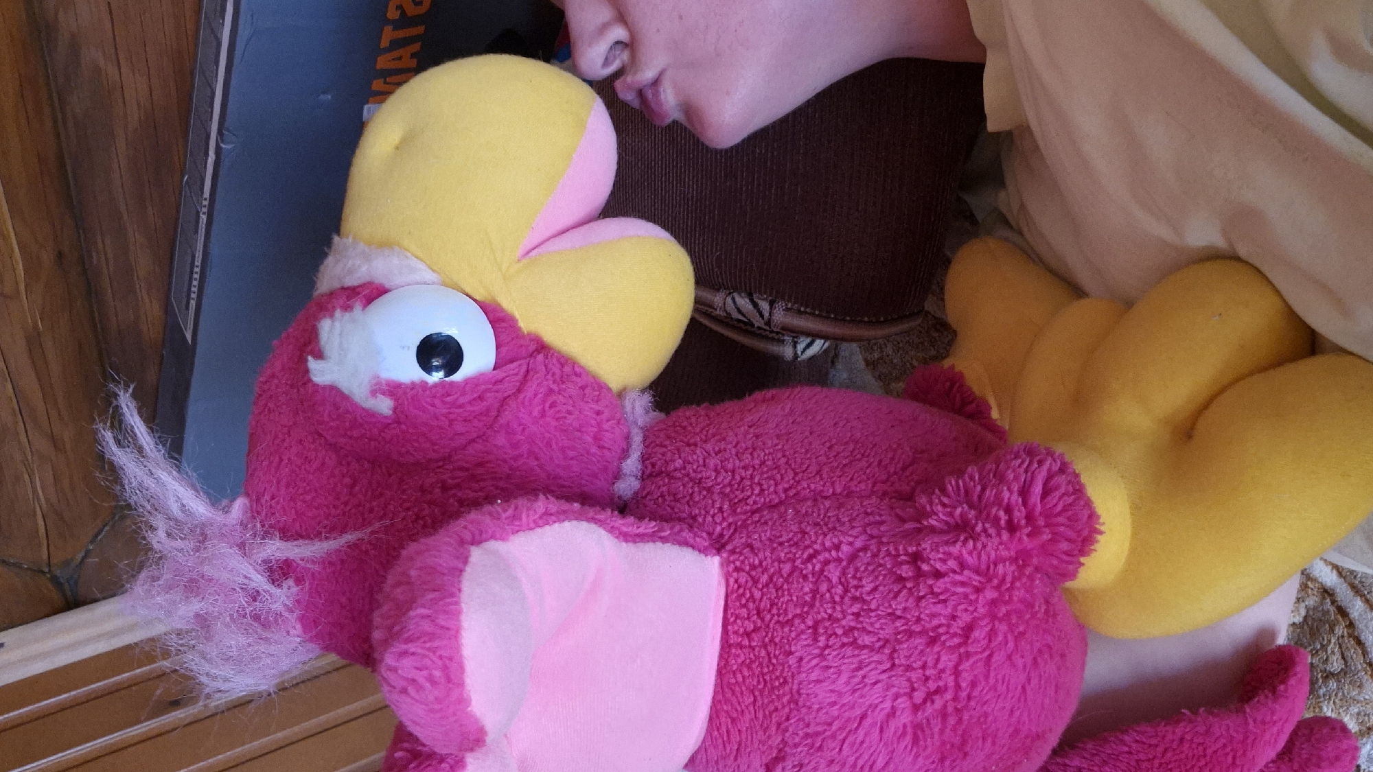 The image depicts a close-up, indoor scene in Новостройка, Russia, focusing on a large, magenta-pink plush toy and the partial profile of a human face.
The central subject is a voluminous, fluffy plush toy, predominantly magenta-pink, resembling a cartoon bird. It features a bright yellow beak with a pink underside, a prominent white eye with a black pupil on its right side, and a sparse tuft of lighter pink, wispy material protruding from the top of its head. The toy's body has pink wing-like appendages on its sides and two yellow, bent leg-like structures at its base.
To the right of the plush toy, the right profile of a human face is visible, encompassing the chin, pursed lips, and a portion of the nose. The skin tone is fair. The human's mouth is positioned directly adjacent to the plush toy's yellow beak, indicating an interaction such as a kiss or nuzzle.
The background elements include light-colored, patterned fabric, likely bedding, in the lower right and foreground. In the upper left and background, wooden paneling or furniture is visible with a light vertical stripe and horizontal wood grain. A dark, rectangular object, possibly a box or furniture piece, with faint white markings and partial text (e.g., "AT?") is present in the upper right background. A dark brown object, appearing to be a cushion or bag, is situated behind the human's head. The scene is evenly lit.