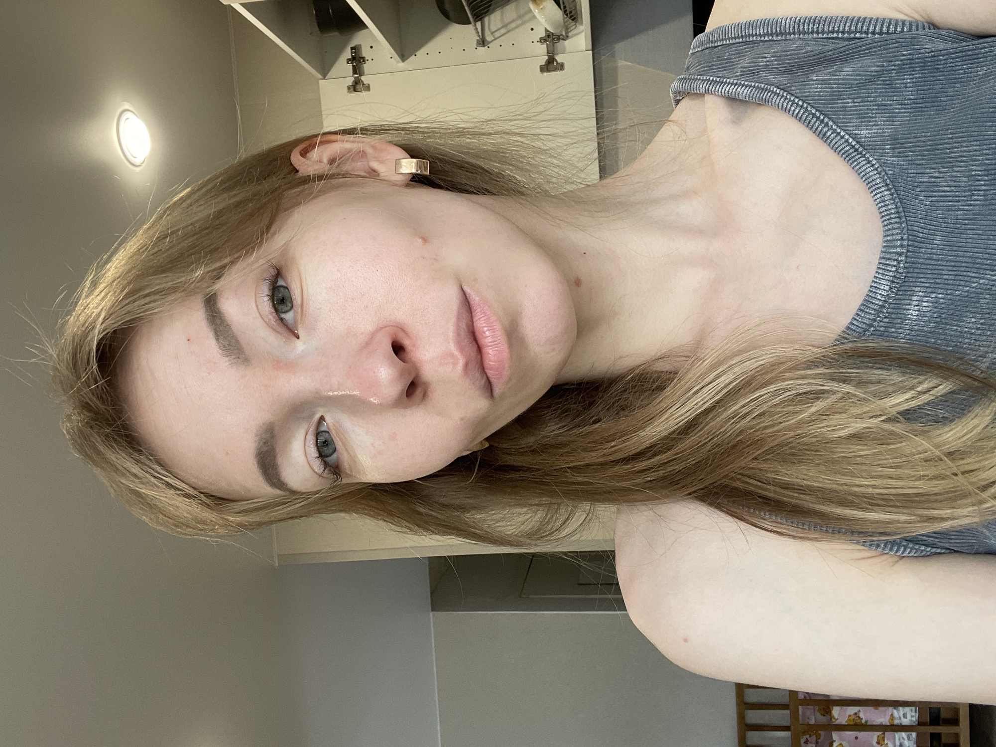 A close-up, angled shot of a young woman with fair skin and light brown hair. She is wearing a gray tank top. She has light eyebrows and eyelashes, and her eyes are a grayish-blue color. A gold, rectangular earring is visible in her left ear. The background shows a kitchen area with white cabinets and a light fixture. The environment suggests an indoor setting, possibly a private residence, in a location with light-colored walls. There is nothing to suggest that the image was taken in Perm, Russia.