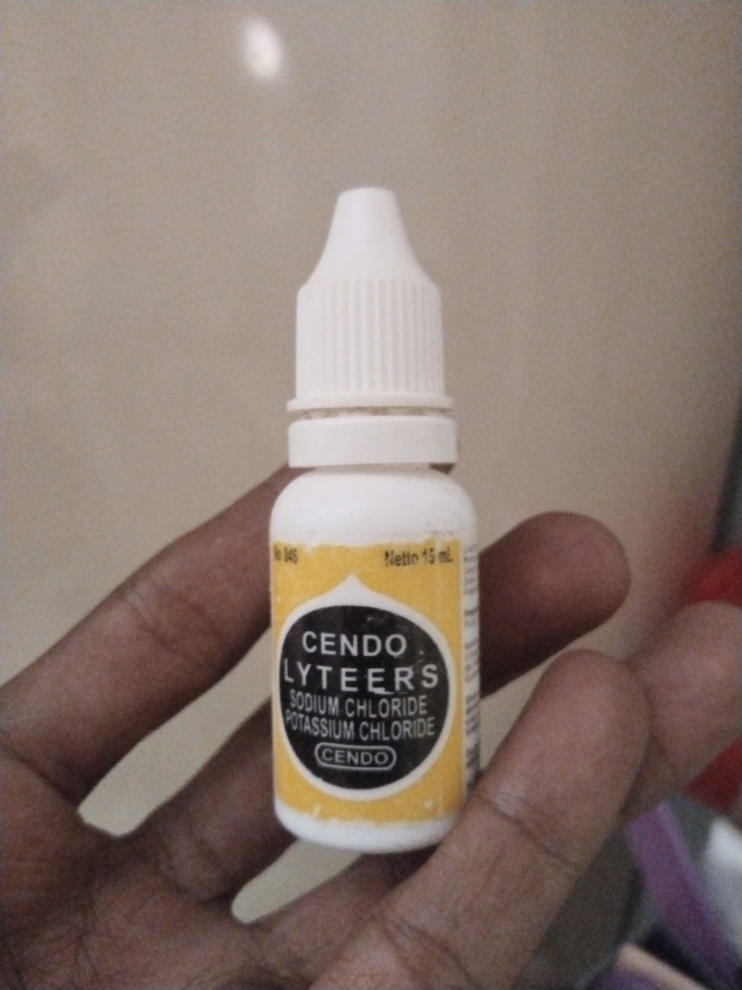 A person's left hand holds a small, white plastic bottle with a white dispensing tip. The bottle has a yellow label with black lettering that reads "CENDO LYTEERS SODIUM CHLORIDE POTASSIUM CHLORIDE CENDO." The label also indicates "Netto 15 mL" and "No 046." The background appears to be a beige wall or surface. It is not possible to confirm the location as Kutaglumpang, Indonesia from the image.