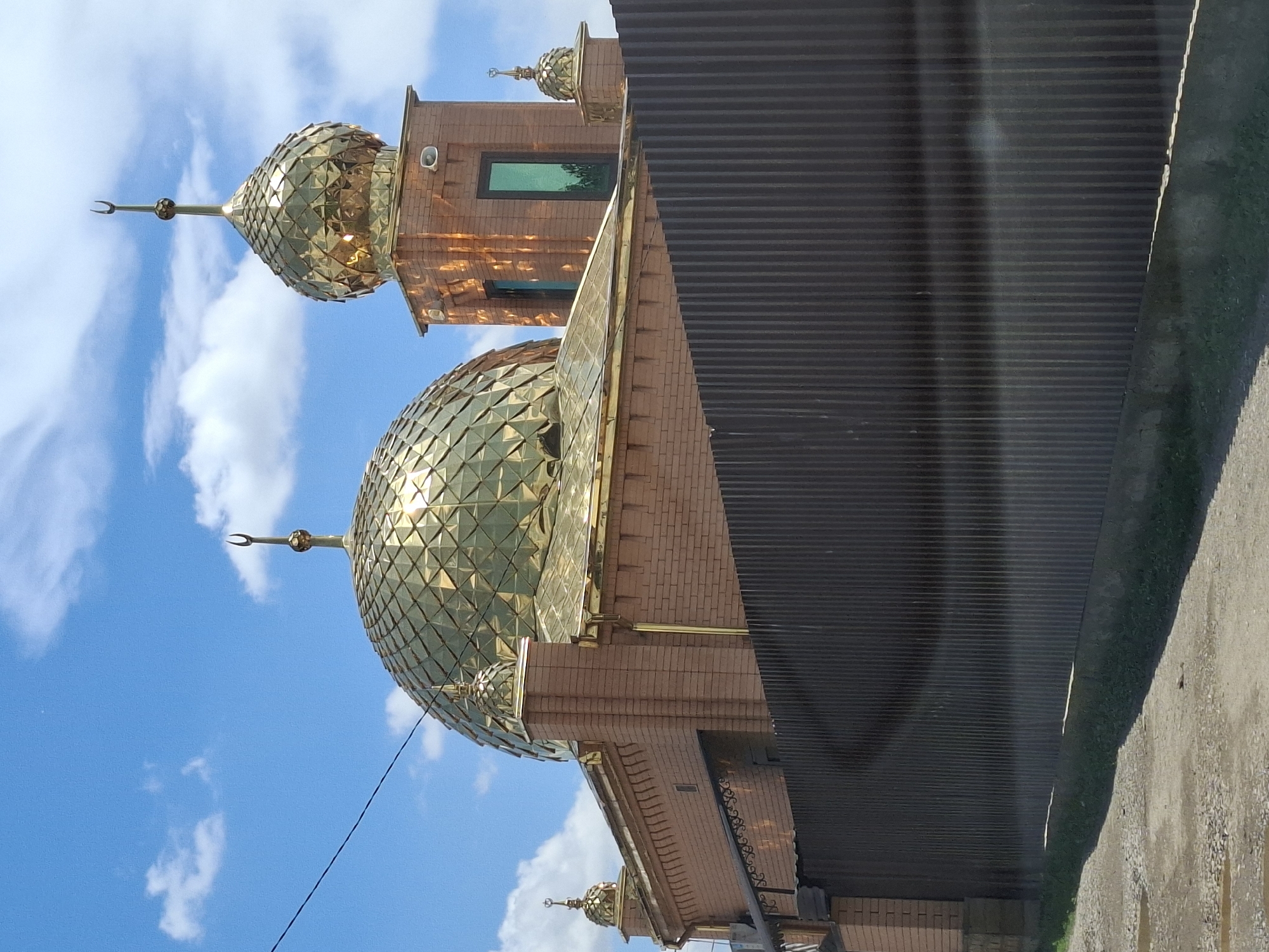 The image shows the exterior of a mosque, likely in Urus-Martan, Russia. The building features prominent golden domes with a faceted surface, reflecting the sunlight. The walls are constructed from reddish-brown brick. A tall, narrow green window is visible on one of the building's sections. The mosque is partially obscured by a dark, corrugated metal fence or wall in the foreground. The sky above is blue with scattered white clouds. A thin black wire is also visible against the sky. The lighting suggests it is daytime, possibly late morning or early afternoon, with bright sunlight casting reflections on the domes. No people are visible in the image. The scene depicts an architectural structure in an outdoor setting.