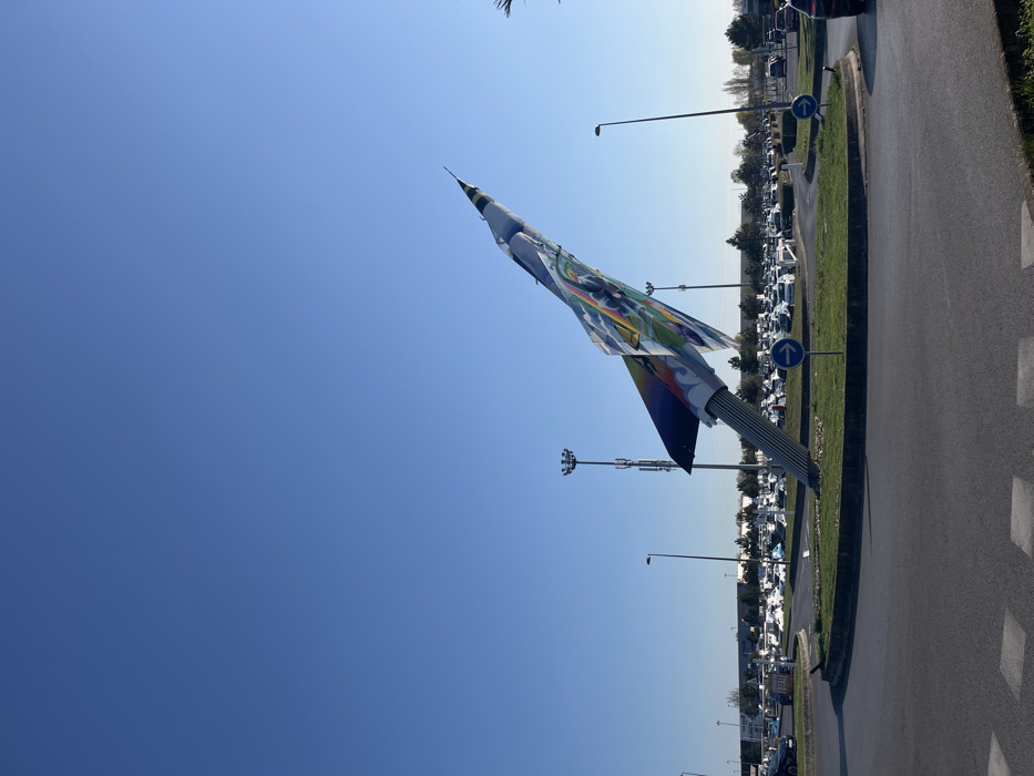 The image shows a large, colorful sculpture of a jet fighter aircraft, painted in bright colors, in a roundabout in Saint-Brice-Courcelles, France. The sculpture is positioned in a grassy area in the center of the roundabout. The roundabout is paved with gray asphalt and surrounded by a curb. There are several cars parked around the perimeter of the roundabout. The sky is a clear blue, and the sun is shining. There are some streetlights and other foliage visible in the background. The sculpture is pointing towards the right side of the image. There are two blue signs with white arrows pointing to the right.