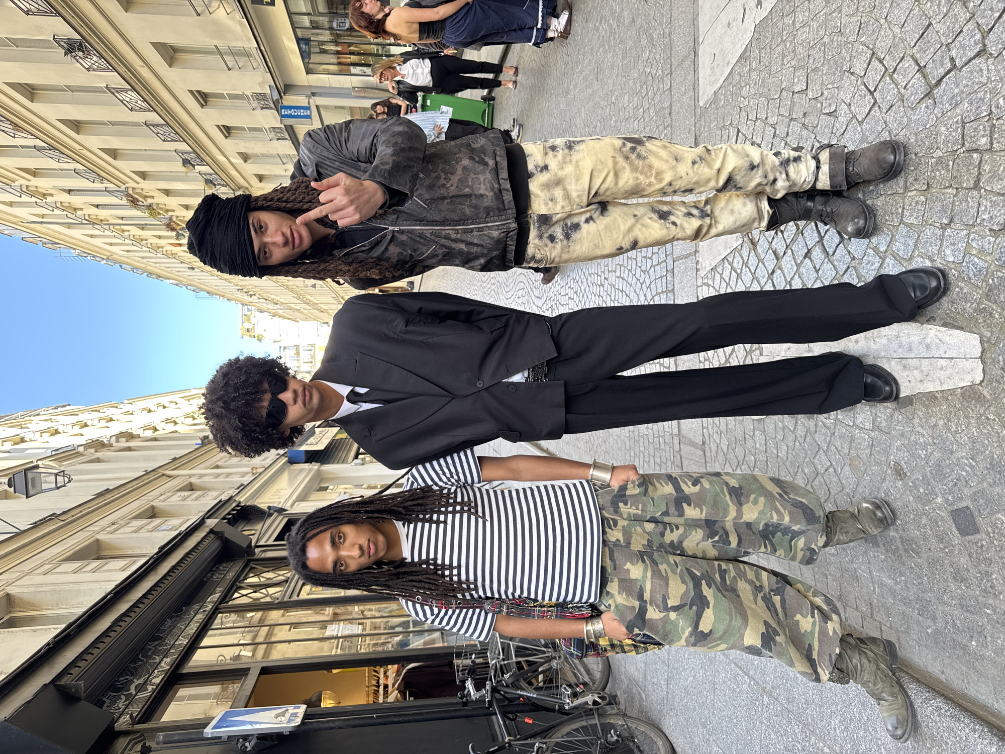 Three young men are captured in a full-length, rotated portrait-style photo on a cobbled street in Paris, France, under a clear blue sky. The image has been rotated 90 degrees clockwise, making the subjects appear to stand horizontally across the frame.
The leftmost subject, with long dreadlocks partially covered by a black head wrap, wears a dark, possibly patterned, zippered jacket over a dark shirt and light-colored, tie-dye or distressed pants, completing his look with dark boots. He looks towards the camera with a subtle smirk and makes a "shaka" hand gesture.
In the middle, another young man with dark, curly hair wears dark sunglasses, a black suit jacket over a white shirt, black trousers, and a visible metallic belt buckle. He also faces the camera directly.
The rightmost subject, featuring long dreadlocks, is dressed in a horizontal black and white striped short-sleeved t-shirt, baggy camouflage cargo pants, and dark boots. Silver-toned wrist cuffs adorn both of his wrists. He maintains eye contact with the camera.
The setting features typical Parisian architecture with tall, light-colored stone buildings flanking the street, adorned with ornate window railings. The ground is paved with cobblestones. In the background, other pedestrians, shop windows, and a green trash bin are visible. A black bicycle is parked near the rightmost subject. A blue street sign displaying "TABAC" is visible on a building facade in the background, indicating a tobacco shop, a common sight in France. The bright conditions suggest it is daytime, likely sunny. The subjects appear to be posing confidently for the photograph.
