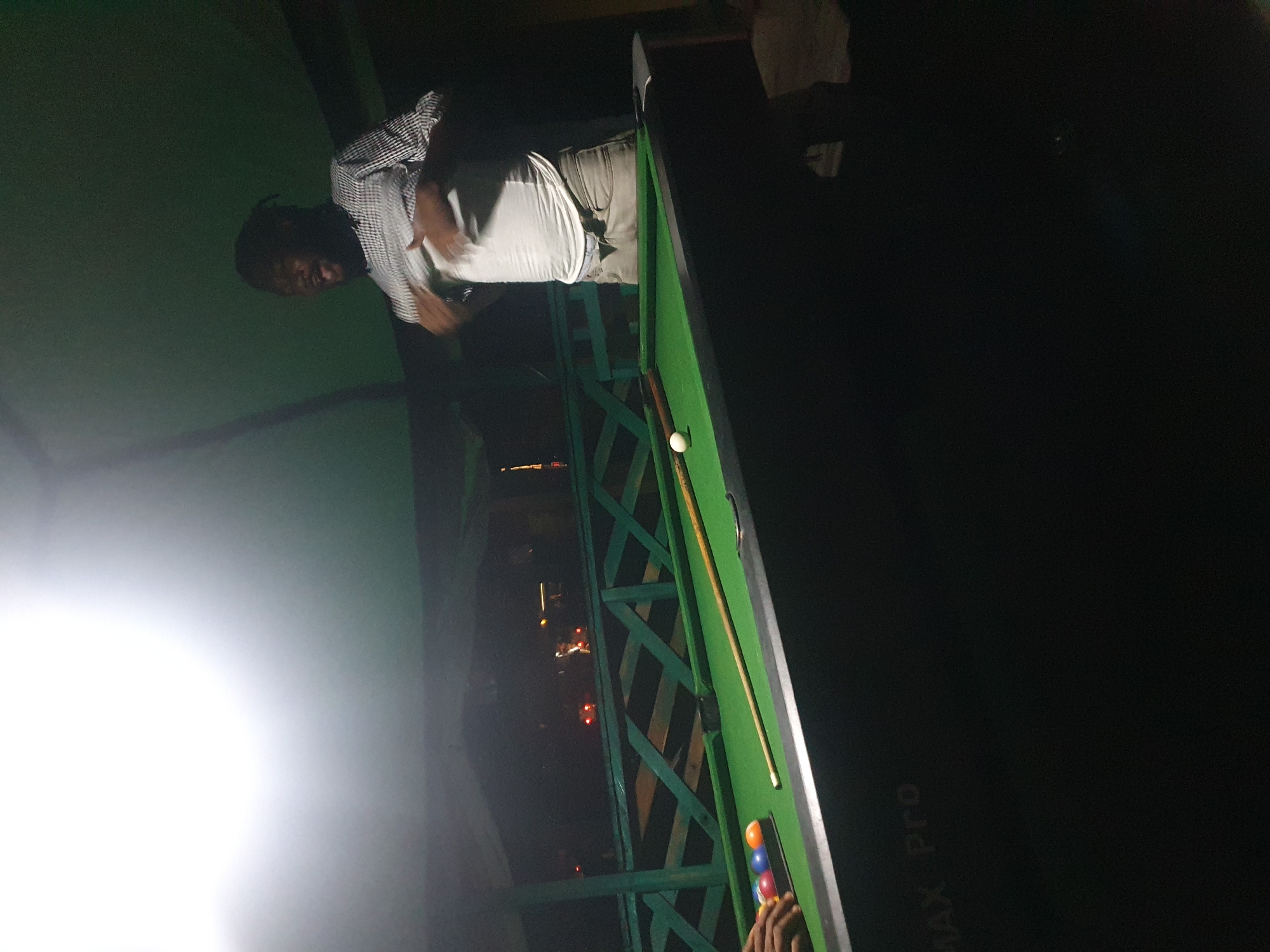 A man in a checkered shirt and light-colored pants is standing next to a green pool table outdoors. He is pulling up his white t-shirt. There are billiard balls and a cue stick on the table. The background shows a wooden fence and a dimly lit cityscape with lights from vehicles. The image appears to be taken at night, possibly in Lokoja, Nigeria, given the setting and the presence of a pool table in an outdoor, possibly recreational, area.