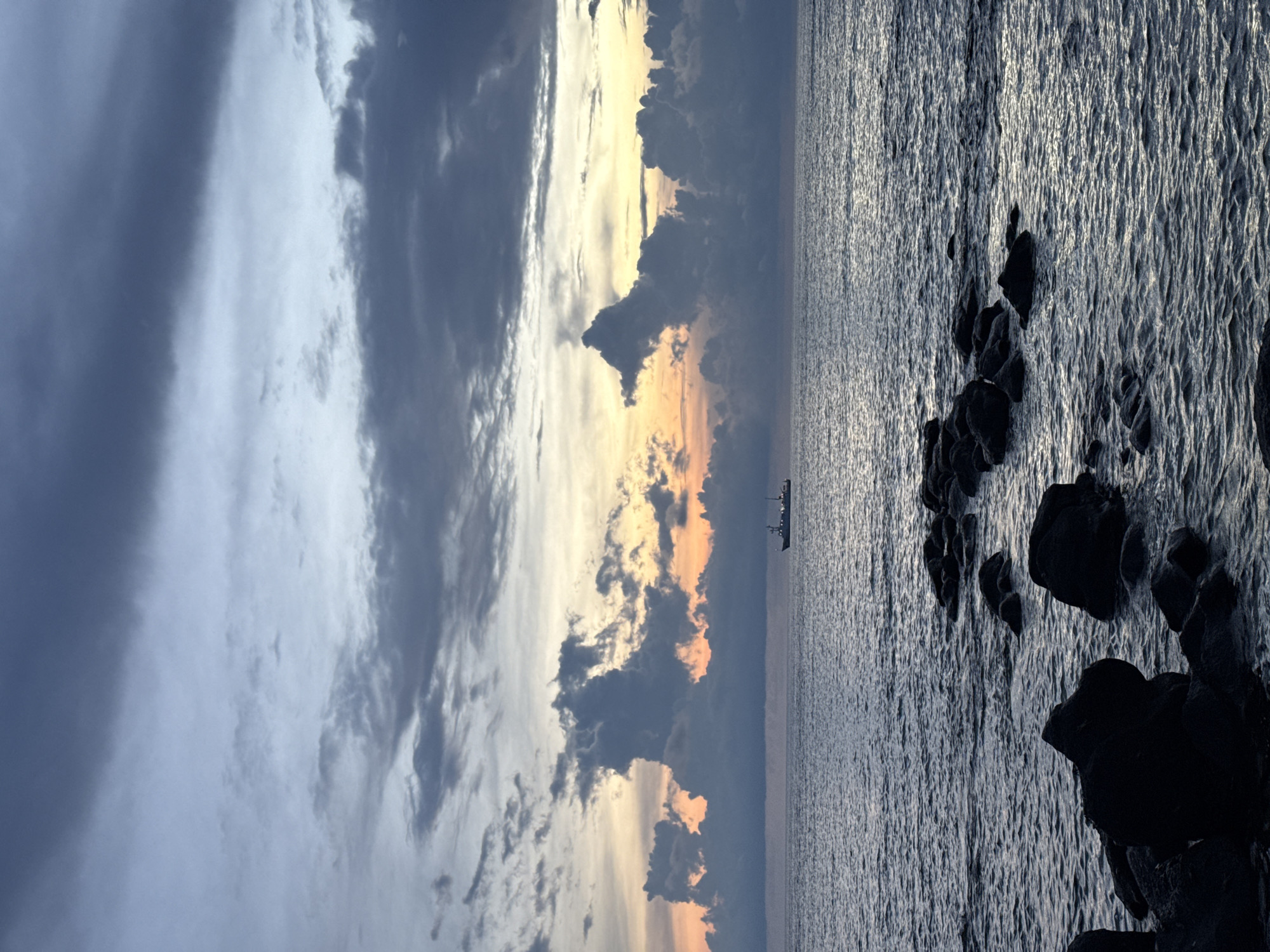 A view of the sea with a ship in the distance, taken from Lý Sơn Special Administrative Region, Vietnam. Dark rocks are visible in the foreground, partially submerged in the water. The sea is calm with gentle ripples. In the middle ground, a ship is sailing on the horizon. The sky is filled with dramatic clouds, with hints of orange and yellow from a sunset peeking through. The clouds are dark and heavy, suggesting an approaching storm or the remnants of one. The overall mood is dramatic and serene.