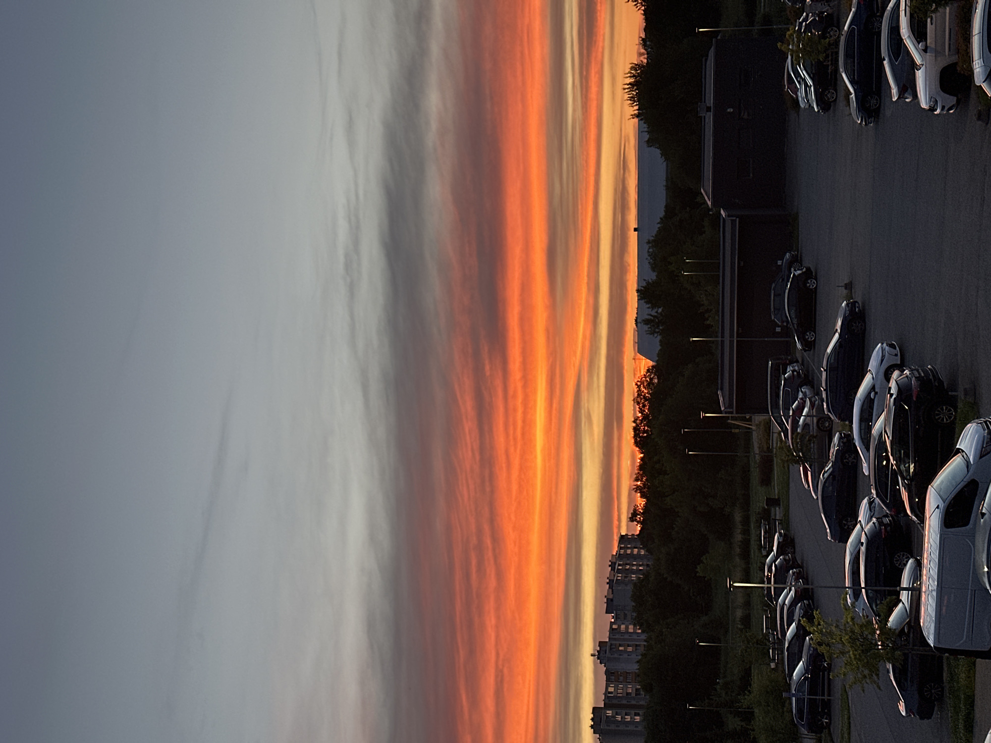 A wide-angle vertical shot captures a sunset over an urban landscape in Tallinn, Estonia. The sky is dominated by vibrant bands of orange and red clouds, transitioning to a pale gray at the top. Below the sky, a line of silhouetted trees separates the horizon from a series of apartment buildings. In the foreground, a parking lot is filled with numerous parked cars of various makes and models. Streetlights dot the parking area. The scene is set at dusk, with the natural light of the sunset casting long shadows and silhouetting objects.