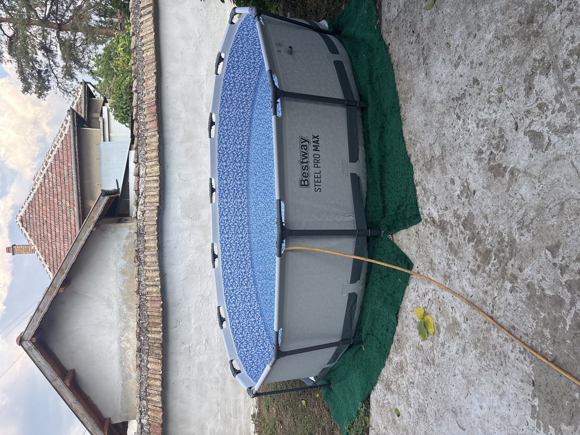 A large, round Bestway Steel Pro Max pool is situated on a green mat in a backyard. The pool is filled with blue water and has a patterned liner. A yellow hose extends from the pool onto a concrete patio. In the background, a white building with a red tile roof and a brick chimney is visible, along with trees and a cloudy sky. The scene appears to be in Brenitsa, Bulgaria.