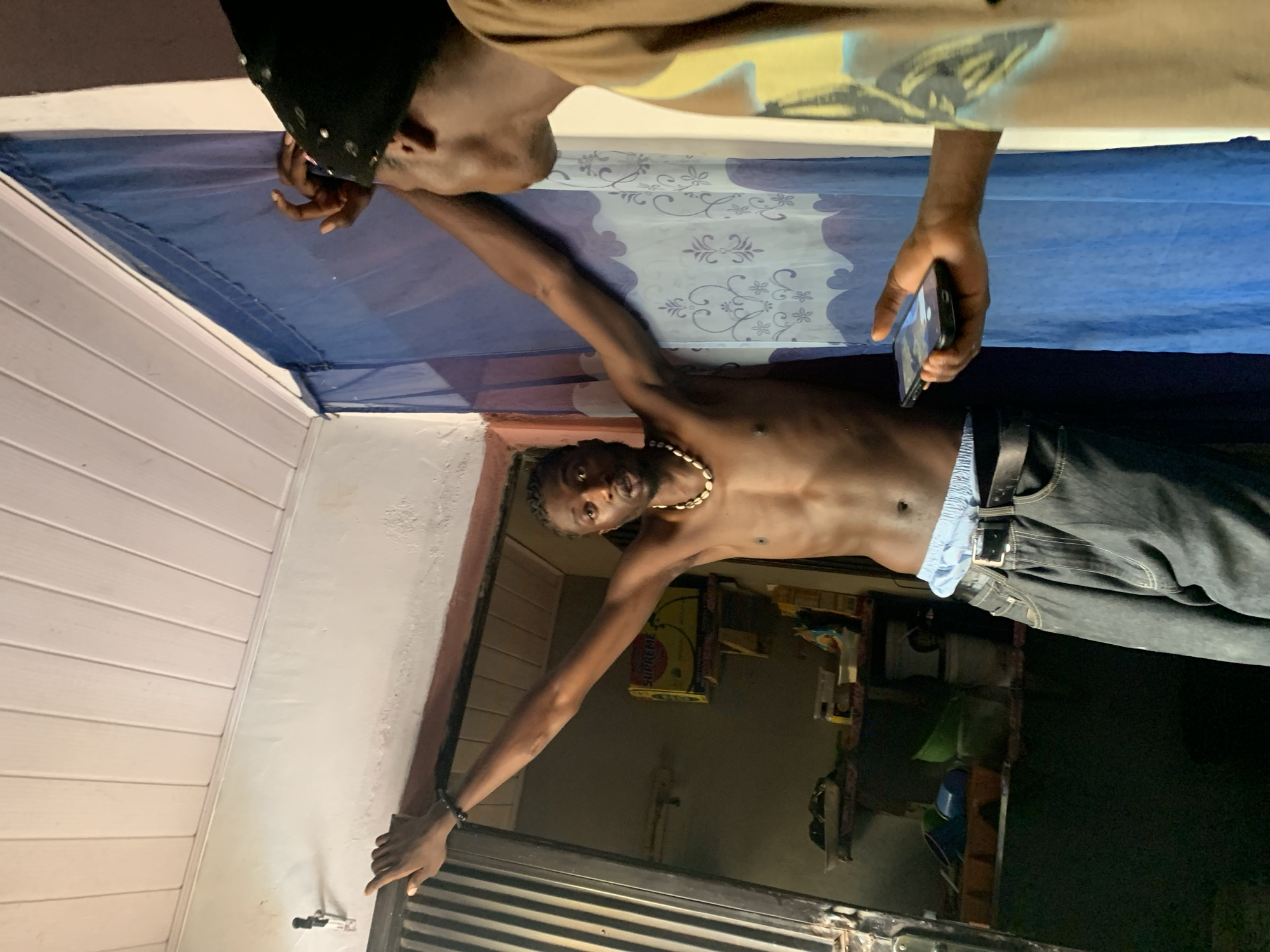 Two young Black males are inside a dimly lit room, possibly a residential dwelling in Agwala, Nigeria. The male in the foreground, facing away from the camera, wears a black skullcap with pearl-like embellishments and a tan t-shirt. He holds a smartphone in his right hand, angled towards the other individual. The second male, shirtless and facing the camera, stands with his arms outstretched, leaning against a blue sheer curtain patterned with floral motifs. He wears blue boxer briefs, a black belt, and dark wash jeans. A shell necklace is visible around his neck, and a beaded bracelet adorns his left wrist. The background reveals shelving units stocked with various items, including boxes and containers. A metal-framed door with vertical slats is partially visible to the left. The room's ceiling is paneled with light-colored wood.