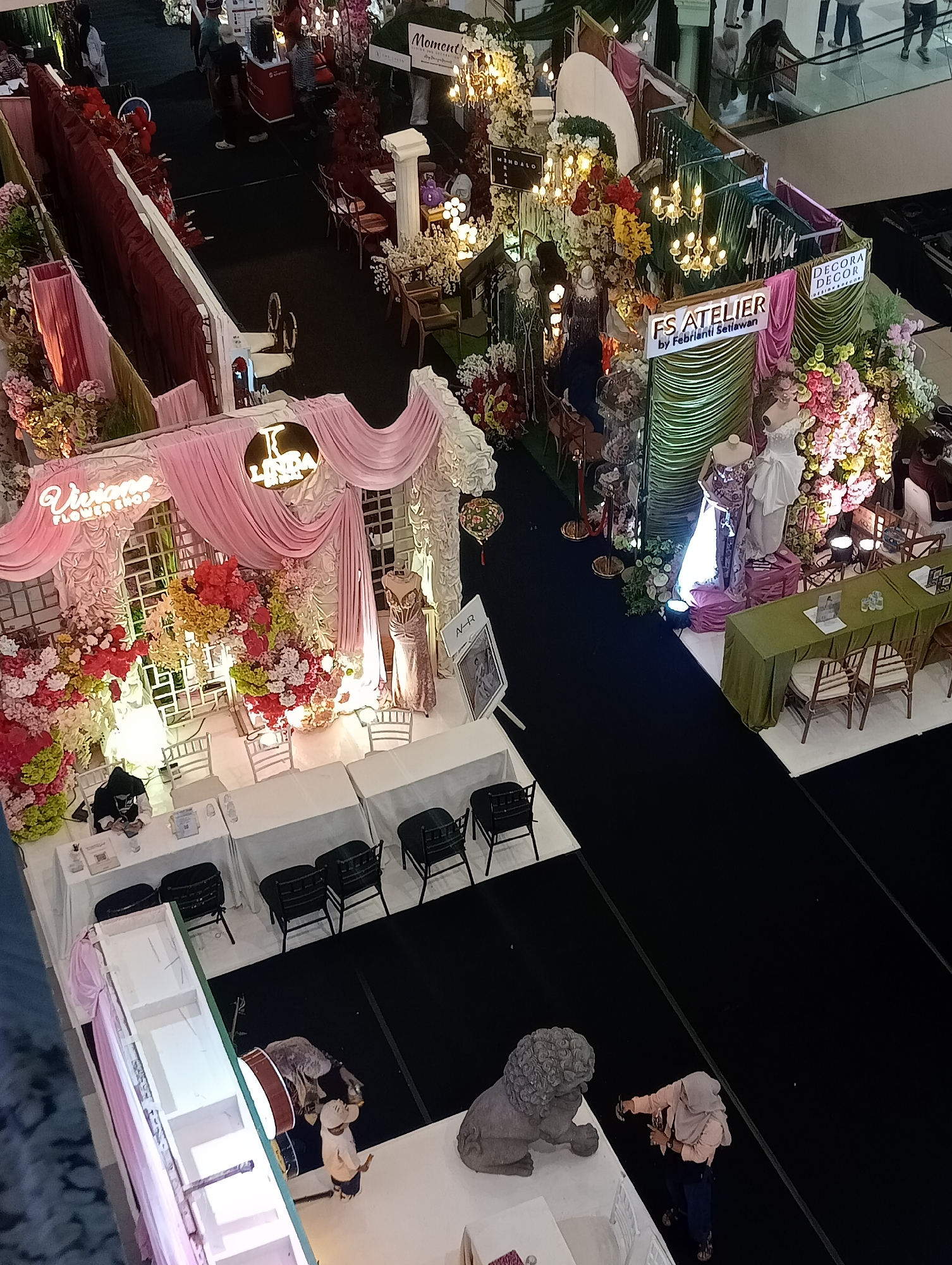 This is a high-angle, full shot of a wedding exhibition, likely held in a mall in Jambi, Indonesia. The scene is dominated by elaborate floral displays and decorative booths.
In the foreground, a booth for "Viviano Flower Shop" is prominently featured, with pink drapes, white lattice work, and abundant red, yellow, and white flowers. Two mannequins are dressed in bridal gowns at another booth labeled "FS ATELIER by Febriani Setiawan," which is adorned with green drapes and further floral arrangements. Another booth displays a sign for "DECORA DECOR." Several white tables and chairs are set up, suggesting a reception or seating area.
In the background, people are seen browsing the exhibition, with some individuals interacting with the displays. Escalators and other mall architecture are visible in the upper portion of the image. A decorative lion statue is present on the right side of the scene, near a woman wearing a headscarf and holding a phone. The overall atmosphere is festive and opulent, indicative of a wedding-related event.