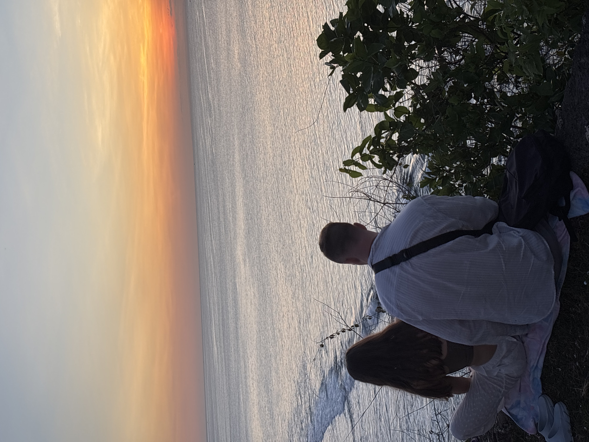 A couple is sitting on a cliff overlooking the ocean at sunset in Kuta Selatan, Indonesia. A man in a striped shirt is next to a woman with long brown hair. They are sitting on a blanket near a tree, facing the sea.