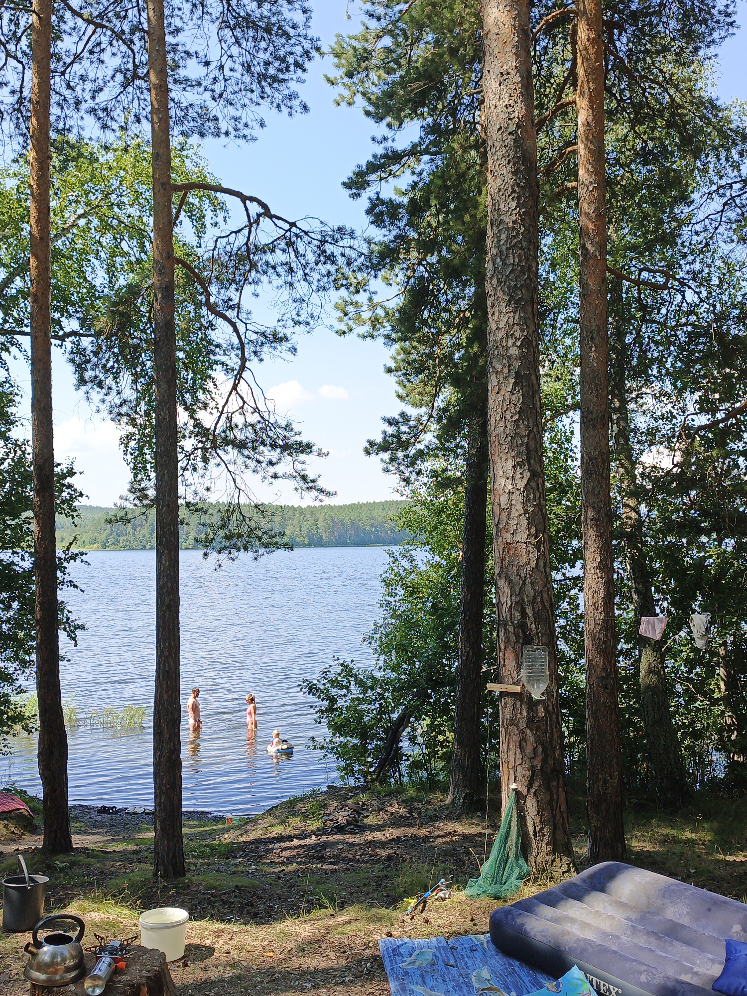 This is a medium-shot, eye-level, outdoor, vertical photograph taken at a lake in Nepryakhinskoye Sel'skoye Poseleniye, Russia. In the foreground, there are several objects on the ground including a camping stove, a kettle, a bucket, and an inflatable mattress. In the middle ground, three people are in the water, two standing and one on a float. The background features a body of water and a dense forest. The lighting is natural and bright, suggesting it is daytime. The overall scene depicts a recreational setting at a lake.