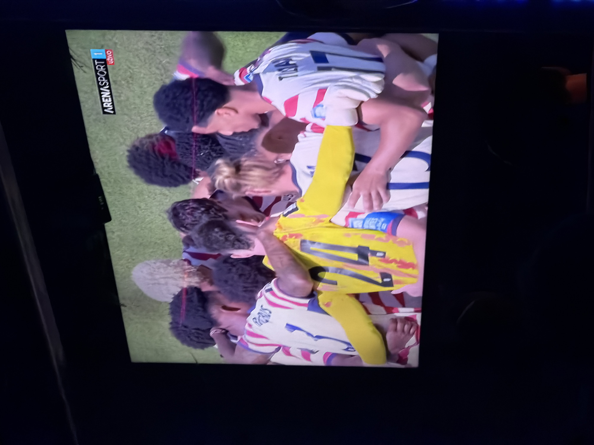 The image shows a screen displaying a sports broadcast. The broadcast features a group of soccer players in a huddle, wearing white jerseys with red and blue accents. One player in the foreground is wearing a yellow jersey with the number 24 on the back. The broadcast logo in the upper right corner reads "ARENASPORT 1 UŽIVO". The screen is positioned in a dark room, likely a home or bar, with the bottom of the screen obscured by darkness. There is no visible information in the image that would place it in Travnik, Bosnia and Herzegovina.