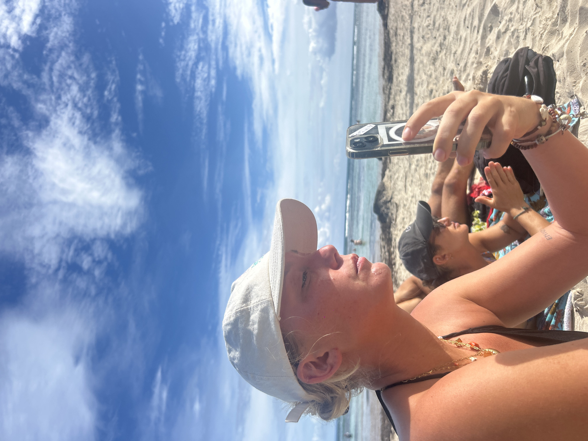 A person is holding a smartphone up in front of them at a beach. The person is wearing a white baseball cap, a necklace, and has bracelets on their wrist. Another person is in the background. The sky is blue with some clouds, and the ocean is in the background as well. It appears to be a sunny day at the beach. The location is unknown.