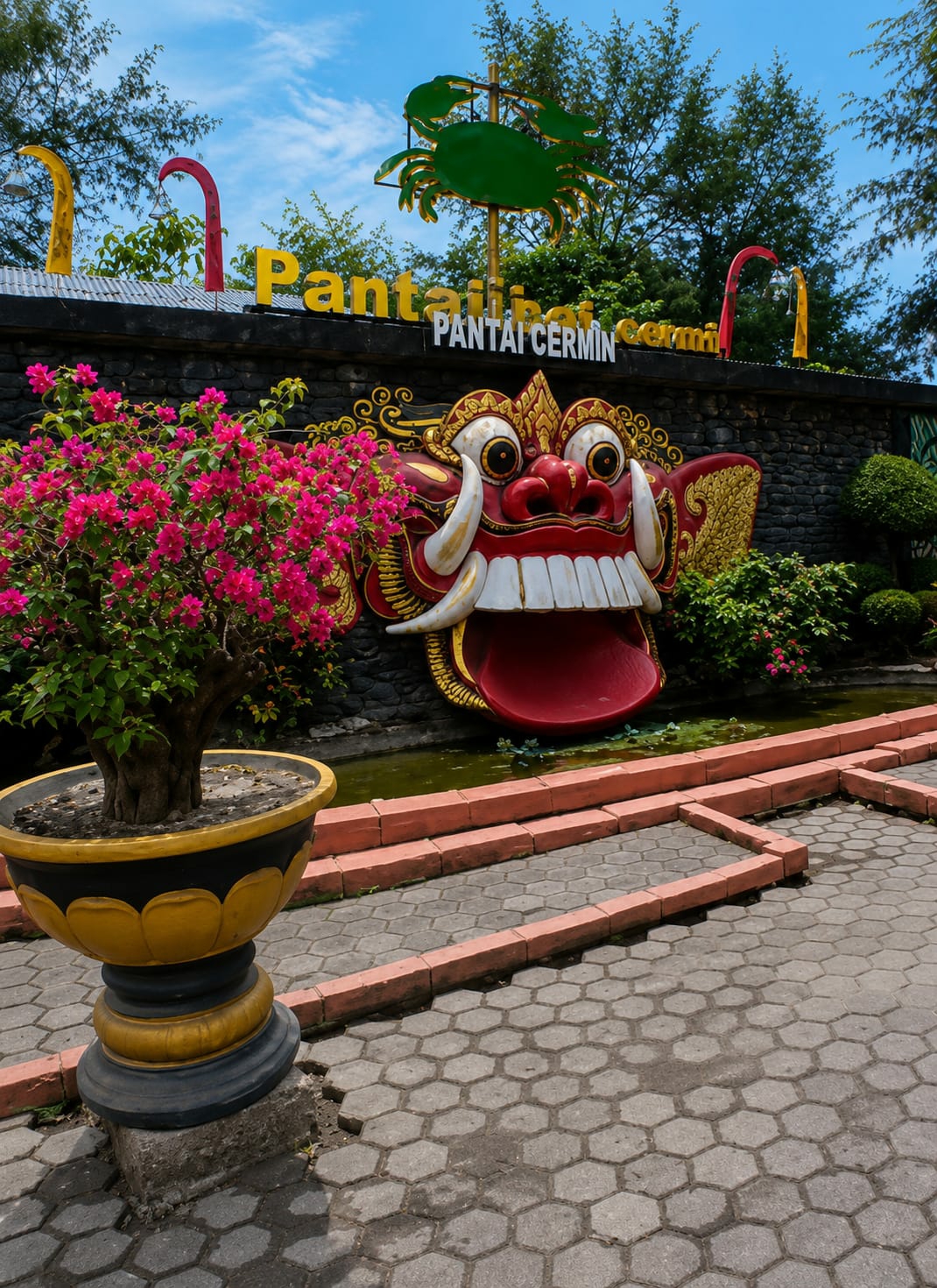 In the city of Kota Pari, Indonesia, a vibrant and slightly unsettling scene unfolds. Dominating the center of the image is a large, grotesque mask, painted in vivid reds, yellows, and whites. Its wide, toothy grin and bulging eyes give it a fierce, almost monstrous appearance. The mask is mounted on a dark, stone-like wall, which serves as a backdrop. Above the mask, yellow lettering spells out "Pantai" and "Pantai Cermin" in Indonesian, indicating the location is a beach called Cermin Beach. Flanking the mask on the wall are decorative elements, including stylized golden wings and red and yellow poles. In front of the wall, a small pond with green water is visible, bordered by coral-colored brick steps. To the left of the mask, a potted plant with abundant pink bougainvillea flowers adds a splash of color and softness to the otherwise intense imagery. The plant sits in a large, decorative black and yellow planter. The foreground is paved with hexagonal grey stones. The sky above is a clear blue with scattered white clouds. The overall impression is one of a tropical, possibly tourist-oriented location with a unique, culturally inspired artistic expression.