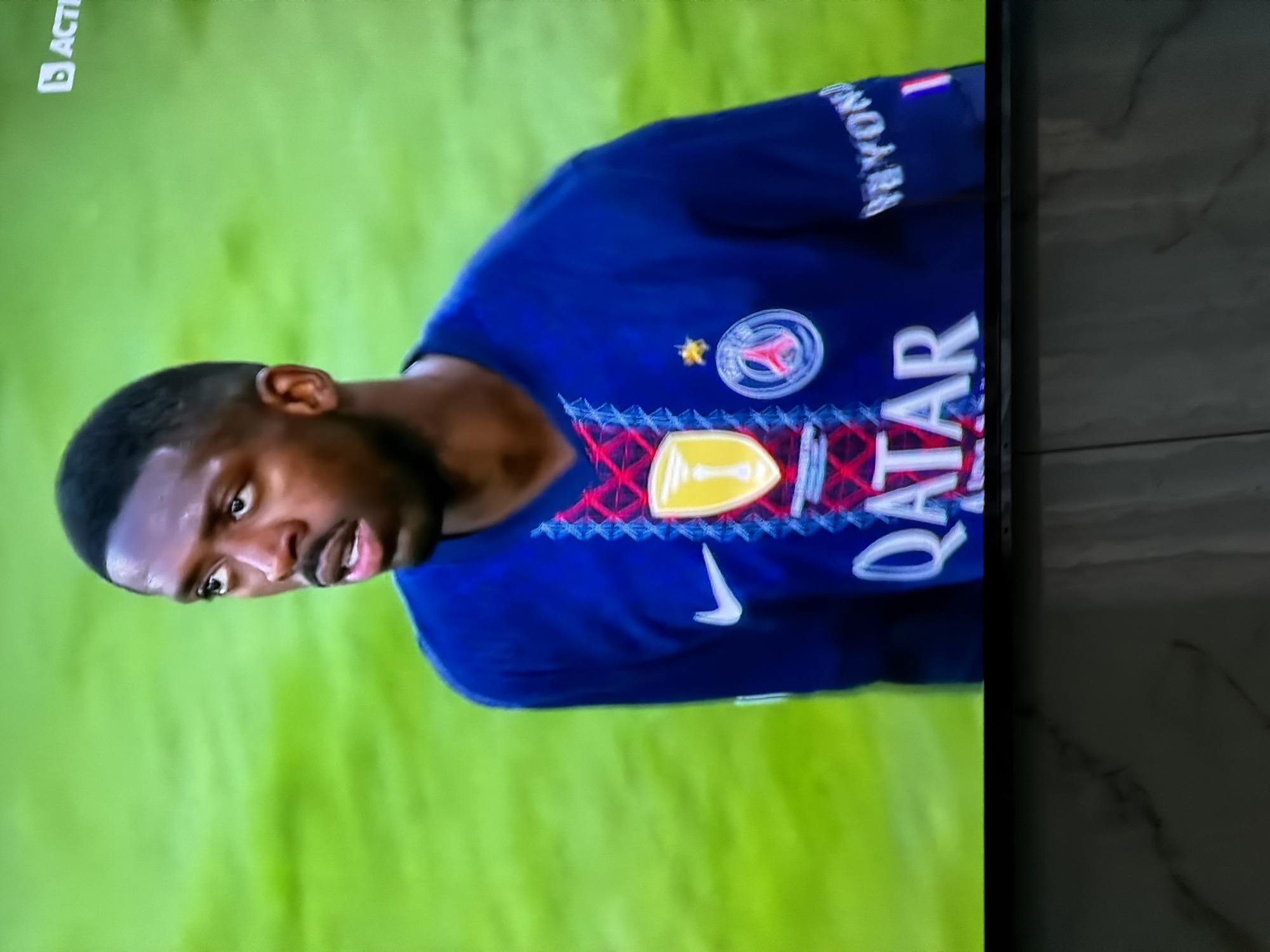 The image shows a man wearing a blue Paris Saint-Germain football jersey with the word "QATAR" on the front. The Nike logo is also visible on the jersey. The background appears to be a blurry green field. The image was likely taken on a television screen. There is nothing in the picture to indicate that the picture was taken in Asenovgrad, Bulgaria.
