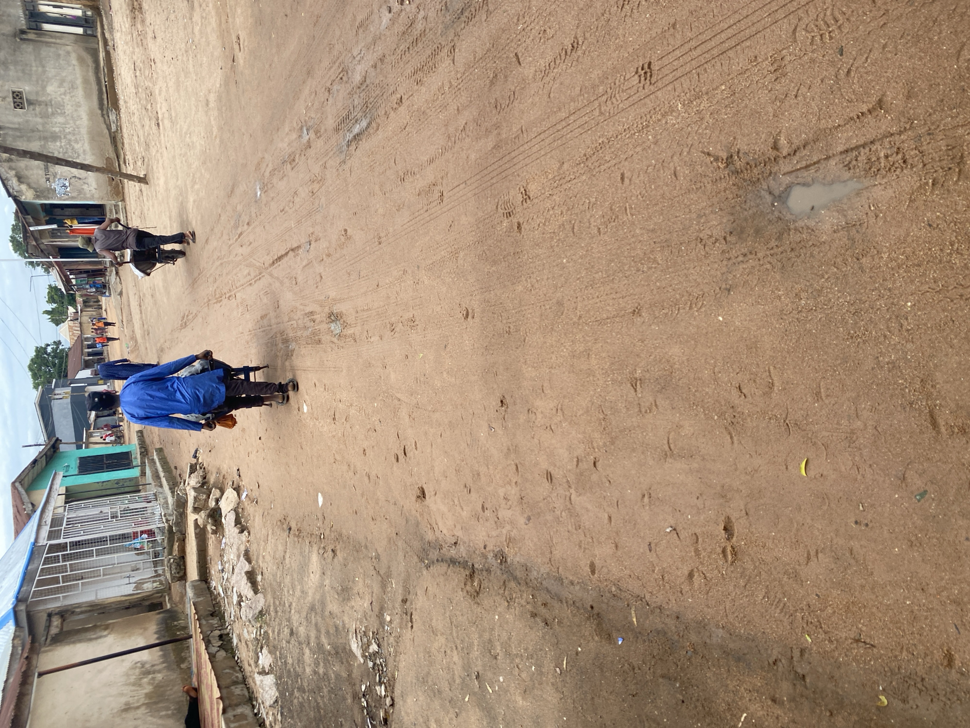 A person wearing a blue shirt and dark pants is riding a motorbike on a dirt road. The motorbike has a blue frame and a yellow bag attached to the back. The person is wearing dark sandals. In the background, there are buildings with corrugated metal roofs and barred windows, typical of residential areas in Jalingo, Nigeria. Some of the buildings appear to be shops. The road is dusty and has tire tracks from vehicles. There are some scattered rocks and debris along the side of the road. Another person is visible further down the road, also on a motorbike. The scene appears to be set during the day, with soft lighting suggesting it might be late afternoon. There is no visible text in the image.
