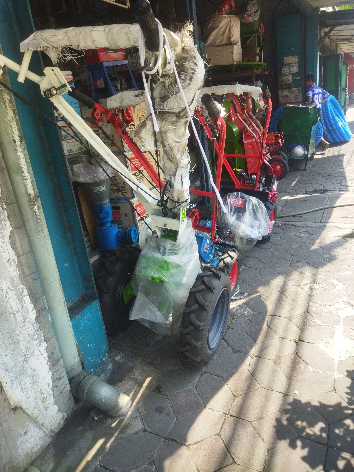 In the city of Giripurwo, Indonesia, on a sunlit paved ground, several pieces of agricultural machinery are displayed outdoors. The scene features two red and green tillers, one with its engine covered in a clear plastic bag. Adjacent to them, a white and blue agricultural machine is parked. In the background, a young person wearing a blue shirt with the number "88" and the word "CHICAGO" is leaning against a green container. Blue plastic tubing is stacked in the container. The overall environment appears to be a market or a storage area for agricultural equipment, with various boxes and materials piled up. A grey downspout is visible on the left side of the image, attached to a wall. The sunlight casts distinct shadows across the patterned pavement.