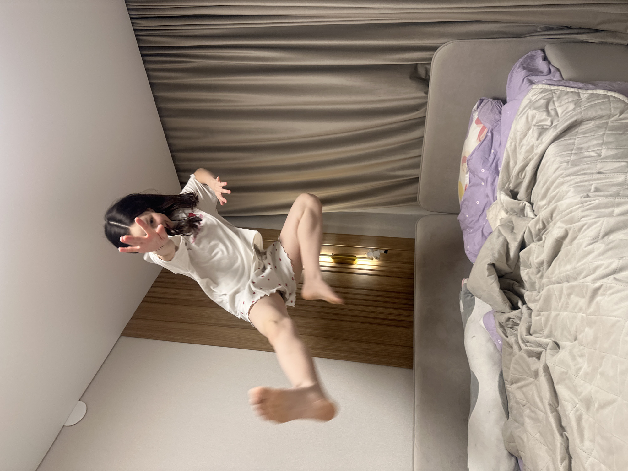 A young girl with long, dark hair is jumping toward the camera. She is wearing a white top with short sleeves and patterned shorts. Her arms are outstretched and her feet are off the ground. The background includes a white wall, a wooden headboard, a beige curtain, and a grey bed with a quilt and patterned sheets. The location is a residential interior, but there is not enough information to specify the city of Votkinsk, Russia.