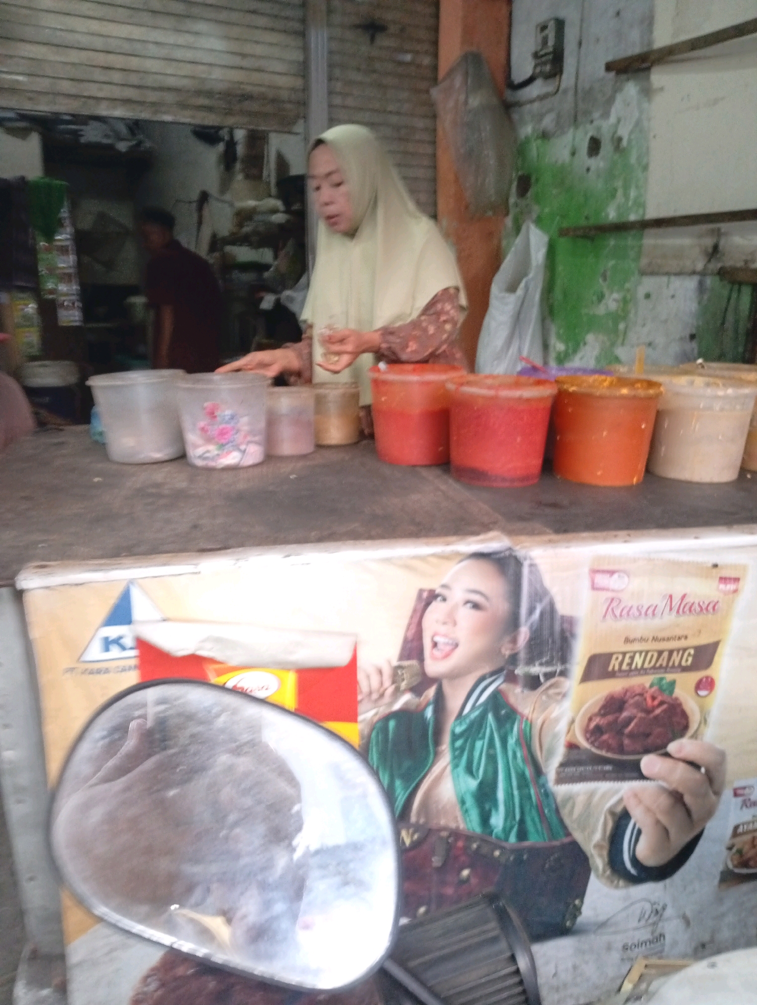 The image shows an outdoor market stall in North Jakarta, Indonesia. A woman wearing a light yellow hijab stands behind a table filled with various containers of food ingredients. To her right, a man in a maroon shirt is partially visible. In the foreground, a large advertisement features a woman holding a packet of "Rasa Masa Rendang" spice mix. The stall appears to be made of simple materials, with a corrugated metal roof and a wooden counter. The environment is typical of a bustling street market, with evidence of commercial activity and food preparation.