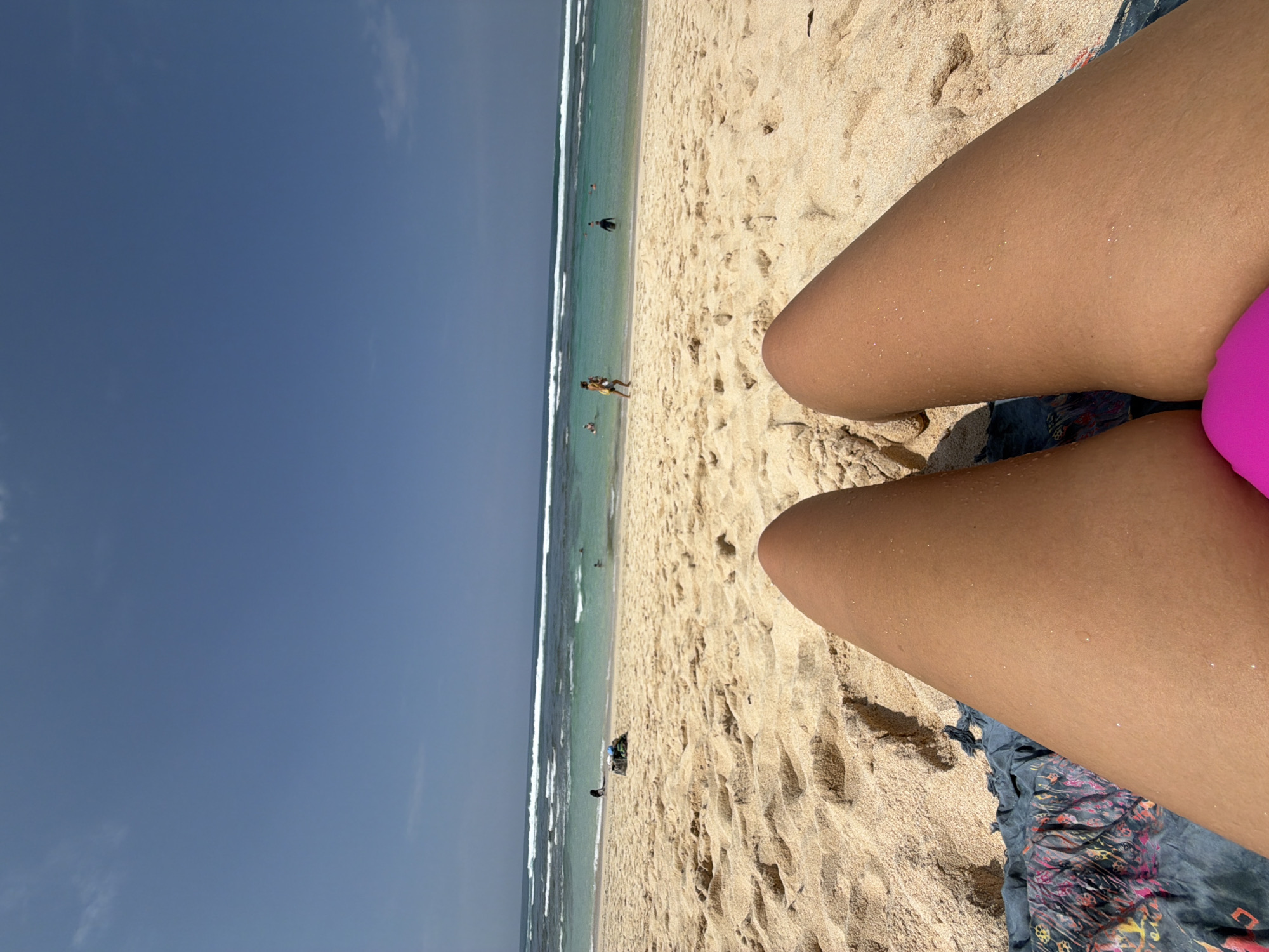 The image displays a first-person perspective of a beach scene in Ungasan, Indonesia. In the immediate foreground, the tanned lower halves of two bare legs are visible, exhibiting water droplets and grains of sand. A section of a bright pink garment is situated between the upper thighs, resting on a dark textile with a colorful, abstract pattern (pink, yellow, blue hues) laid on the sand. The midground features a light-colored sandy beach marked with footprints and minor undulations. A small, dark bundle of items is located on the sand near the water's edge to the left. The turquoise ocean water beyond shows multiple white-capped waves breaking. Several individuals are present in the water: one person in dark attire is partially submerged towards the center-right, another in light-colored shorts is running from the water towards the beach, and additional smaller figures are wading or swimming further out. The background consists of a clear, light blue sky with a few sparse, wispy white clouds.