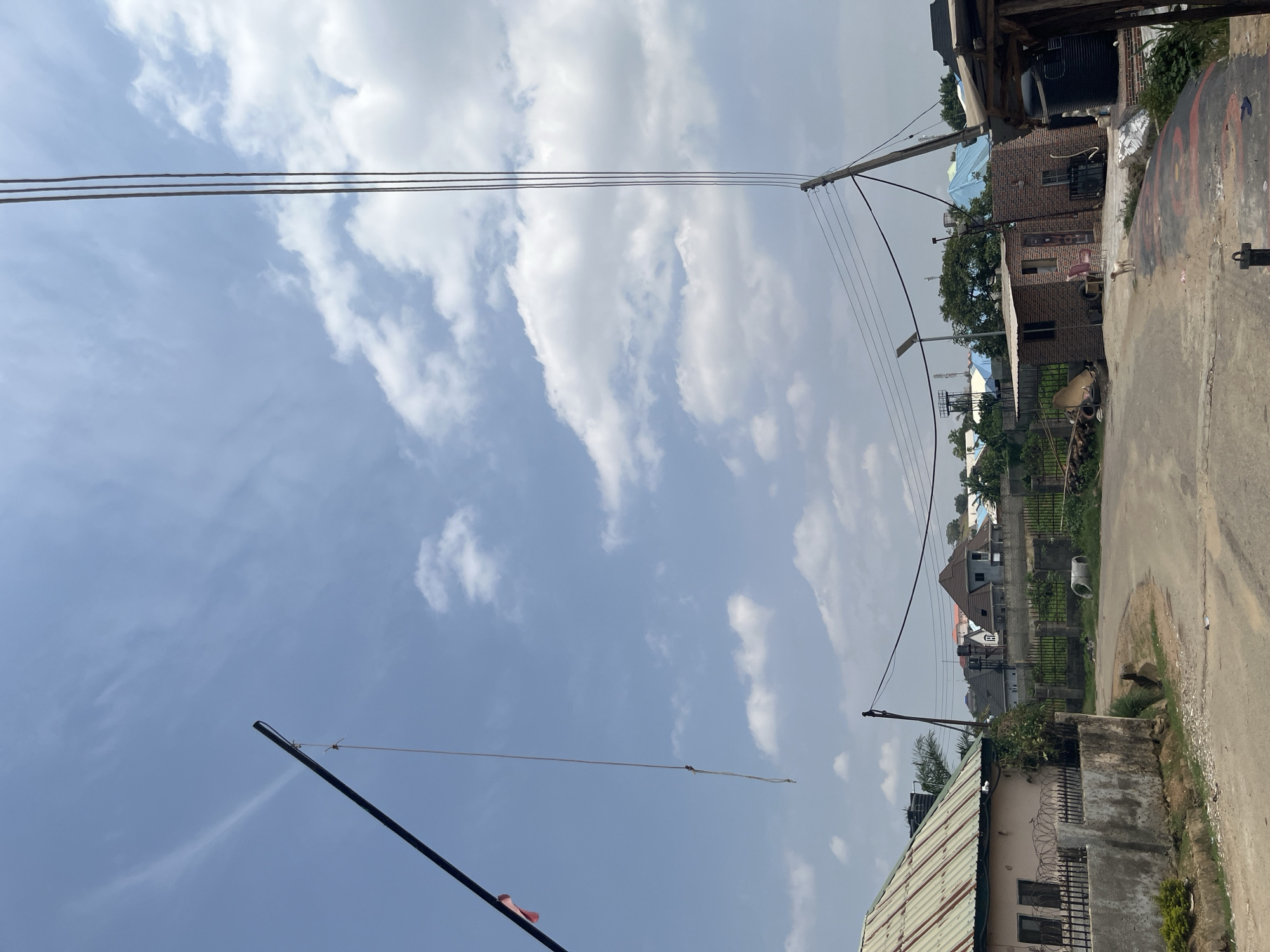 This image depicts a street scene in Abuja, Nigeria. The sky is bright blue with scattered white clouds, suggesting it is daytime, likely mid-morning or early afternoon. Overhead, power lines stretch across the frame, supported by utility poles.
The scene includes several buildings with varying architectural styles. Some appear to be residential homes, constructed with brick and featuring pitched roofs. Others seem to be more modest structures. In the foreground, a corrugated metal roof is visible. Beyond the immediate buildings, more houses and greenery can be seen, indicating a suburban or urban residential area.
On the right side of the image, a paved road runs alongside a patch of unkempt ground. A concrete wall with barbed wire atop it borders the road. There are some scattered objects on the ground, including what appear to be tires. A small animal, possibly a dog, is visible near the wall. Further down the road, other buildings and trees are present, along with what look like cell towers in the distance.
The overall impression is of an active, lived-in environment, typical of many urban areas in Nigeria. There are no people clearly visible in the frame, but the presence of infrastructure like power lines and buildings suggests human habitation and activity. The weather appears pleasant, with good visibility. No text is discernable in the image.