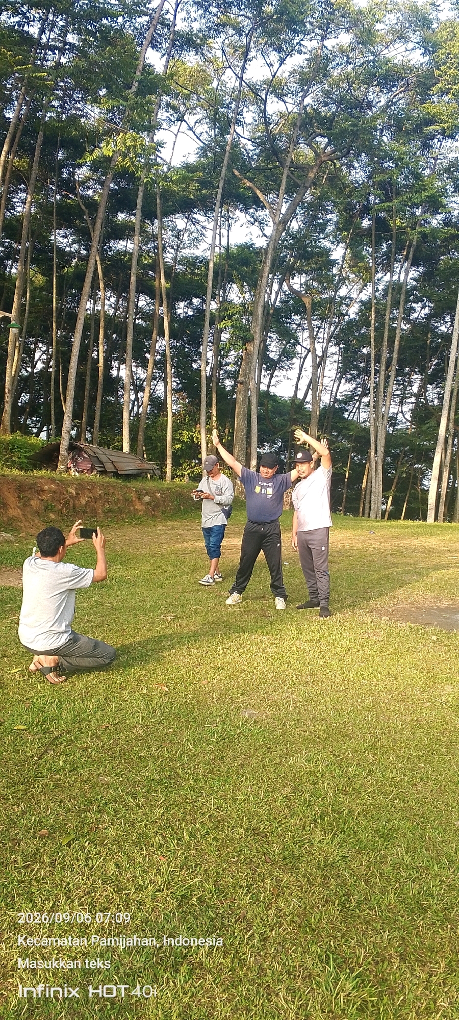 The image depicts an outdoor scene with four individuals amidst a grassy area surrounded by tall trees. In the foreground, a person kneels on the grass, holding a smartphone and appearing to take a photograph. Further back, three other individuals are visible. Two men in the center are standing and performing arm exercises, with one wearing a dark blue t-shirt and black pants, and the other in a light-colored t-shirt and grey pants. Between them and to the left, a third person stands holding a smartphone. The environment is characterized by a well-maintained lawn and a backdrop of lush green trees, suggesting a park or recreational area. The timestamp on the image indicates 2026/09/06 at 07:09, and the location is identified as Pamijahan, Indonesia.