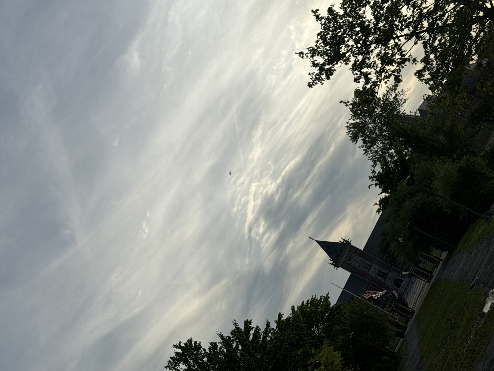 The image captures a slightly angled view of New Rochelle City Hall in New Rochelle, United States of America. The building's clock tower is visible against a sky filled with wispy clouds and contrails of a passing aircraft. Trees frame the top and bottom of the image, partially obscuring the building. An American flag hangs in front of the building. A lamppost stands on the right, beside a paved pathway adjacent to a grassy area.