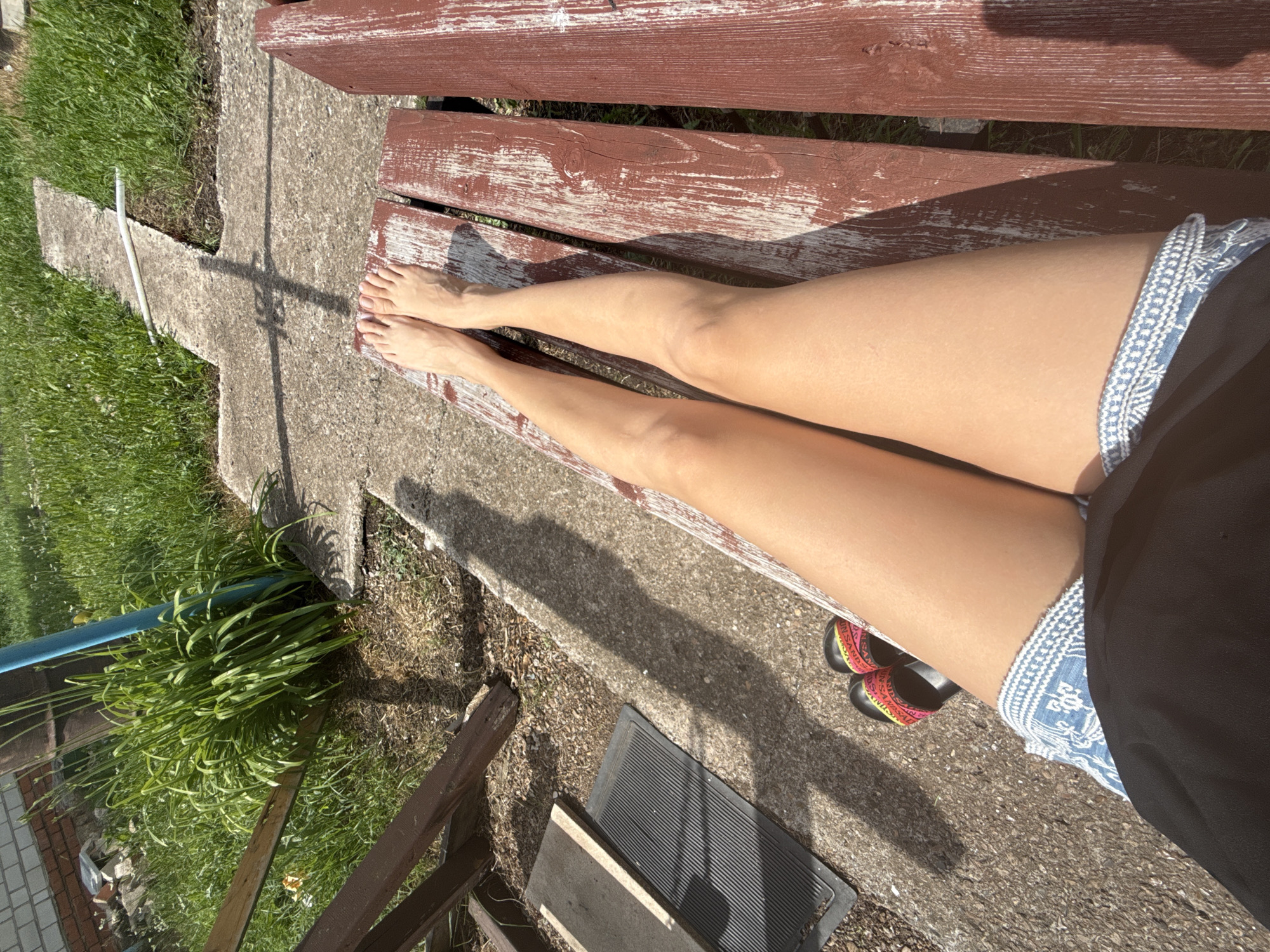 A first-person perspective shot shows a person's legs and feet resting on a weathered, red-brown wooden bench. The person is wearing denim shorts and black sandals with colorful straps. The legs are tanned and appear to be bathed in sunlight, casting a shadow on the concrete ground. The surroundings suggest an outdoor setting, possibly a yard or garden, with green grass, a concrete pathway, and some foliage. A partial view of a brick wall and wooden fencing is visible in the background. The lighting indicates it is daytime, with the sun casting strong shadows.