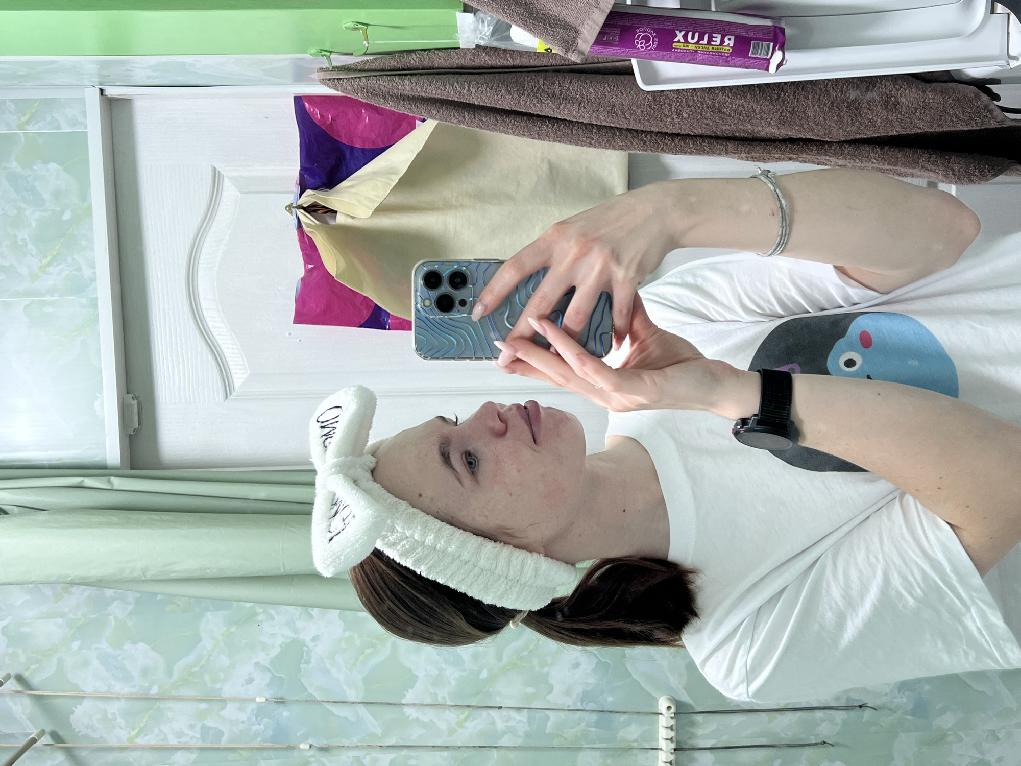 A woman is holding a smartphone in a bathroom setting, likely taking a selfie. She is wearing a white t-shirt with a cartoon graphic, a white fuzzy headband, and a watch on her left wrist. Her hair is brown and pulled back.
The bathroom features a white door and a green and white marble-patterned wall. Above the door are items on a shelf, including a brown towel and a package of "BELUX" cosmetics. A multicolored bag hangs on the door. A clothes drying rack is partially visible at the bottom of the frame. The image was taken indoors. There is no evidence to determine the location in the city of Chaikovsky, Russia.