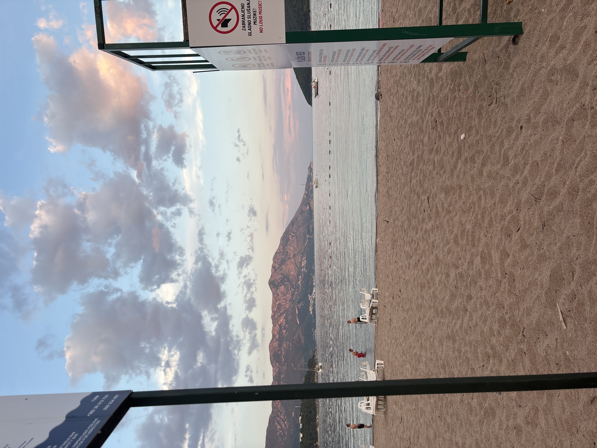 The image captures a beach scene in Budva, Montenegro. The sky features a gradient of blue and pink hues with scattered clouds. The Adriatic Sea stretches towards a mountainous backdrop. The shoreline is marked by a sandy beach dotted with white chairs and a few individuals wading in the water. In the foreground are signage poles with regulatory information, including a 'no loud music' sign, and contact details, with a number reading +382 30 312 733.