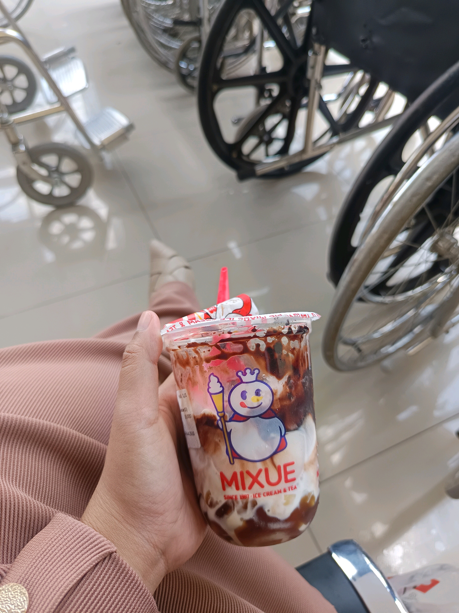 A first-person perspective shot shows a hand holding a plastic cup of Mixue ice cream and tea. The cup is filled with a creamy, layered drink, topped with chocolate syrup and a red straw. The cup has a depiction of a cartoon snowman with a crown. The person is wearing a pink ribbed top. In the background, out of focus, are several wheelchairs, suggesting a hospital or medical facility. The floor appears to be tiled. The overall impression is one of waiting or resting in a public indoor space. There is no specific indication of Bondowoso, Indonesia.