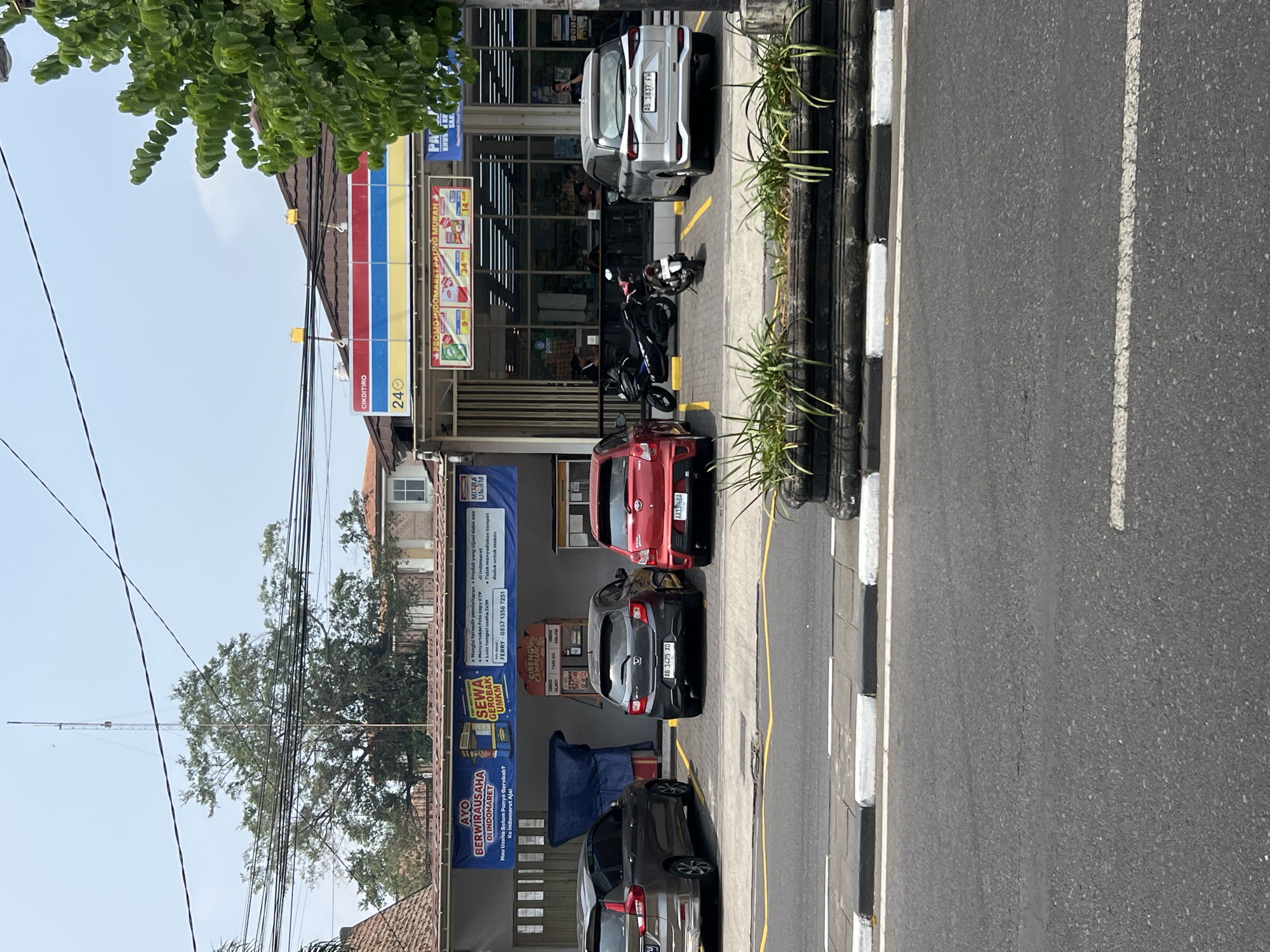 In the city of Yogyakarta, Indonesia, a scene unfolds in front of a minimarket with a distinctive red, yellow, and blue striped awning. The minimarket, identified by the "CIK DITIRO" signage and the "24" indicating its operating hours, features large glass windows revealing an interior with fluorescent lighting. A banner above the entrance promotes "PROMO INDOMARET PALING MURAH," showcasing discounted items.
In the foreground, a row of parked vehicles is visible. From left to right, a dark gray SUV, a dark gray hatchback, a red sedan, and a silver SUV are parked in designated spots marked by yellow lines on the asphalt. Two motorcycles, a black maxi-scooter and a smaller white scooter, are parked behind the red sedan and between the red sedan and the silver SUV, respectively. A dark gray potted planter with green foliage sits on a concrete barrier in front of the parking area, separating it from the paved road.
A wider banner is stretched across the left side of the building, featuring the text "AYO BERWIRAUSAHA DI INDOMARET" (Let's do business at Indomaret) and "SEWA GEROBOK UMKM" (Rent an MSME cart). Smaller text details the benefits of entrepreneurship with Indomaret.
Several individuals are visible within the minimarket, including a person standing near the entrance, possibly an employee, and several customers browsing the aisles. The environment suggests a typical day in a commercial area, with vehicles and pedestrians interacting within the urban landscape. The sky is clear and bright, indicating daylight.
