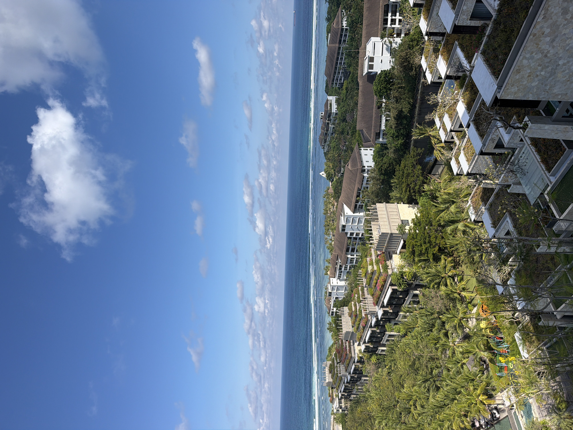 A vibrant high-angle view captures a tropical coastal landscape, showcasing an expansive ocean, a clear blue sky, and a lush resort complex in Kuta Selatan, Indonesia.
The upper portion of the image features a bright blue sky, dotted with scattered white, cumulus clouds of various sizes, indicating sunny and pleasant weather conditions. Below the sky, a wide expanse of deep blue ocean stretches horizontally, its surface showing subtle variations in color. White-capped waves are visible breaking closer to the shore, and a small, dark vessel can be faintly discerned on the distant horizon.
The right side and bottom of the frame reveal a densely developed coastline, home to numerous multi-story resort or hotel buildings. These structures exhibit modern architecture, predominantly with white or light-colored facades and flat or dark-tiled roofs. Many buildings feature terraced designs, balconies, and flat roofs adorned with green plantings or small gardens, seamlessly integrating with the natural environment. The resort complex is enveloped by abundant tropical vegetation, including a multitude of tall palm trees and dense greenery that creates a verdant setting. Nestled among the trees, a colorful children's playground with slides and other equipment is visible, suggesting family-friendly amenities.
The scene is bathed in bright daylight, likely late morning or early afternoon. No specific human activity is clearly visible, but the setting implies leisure, relaxation, and vacation activities. There is no discernible text within the image.