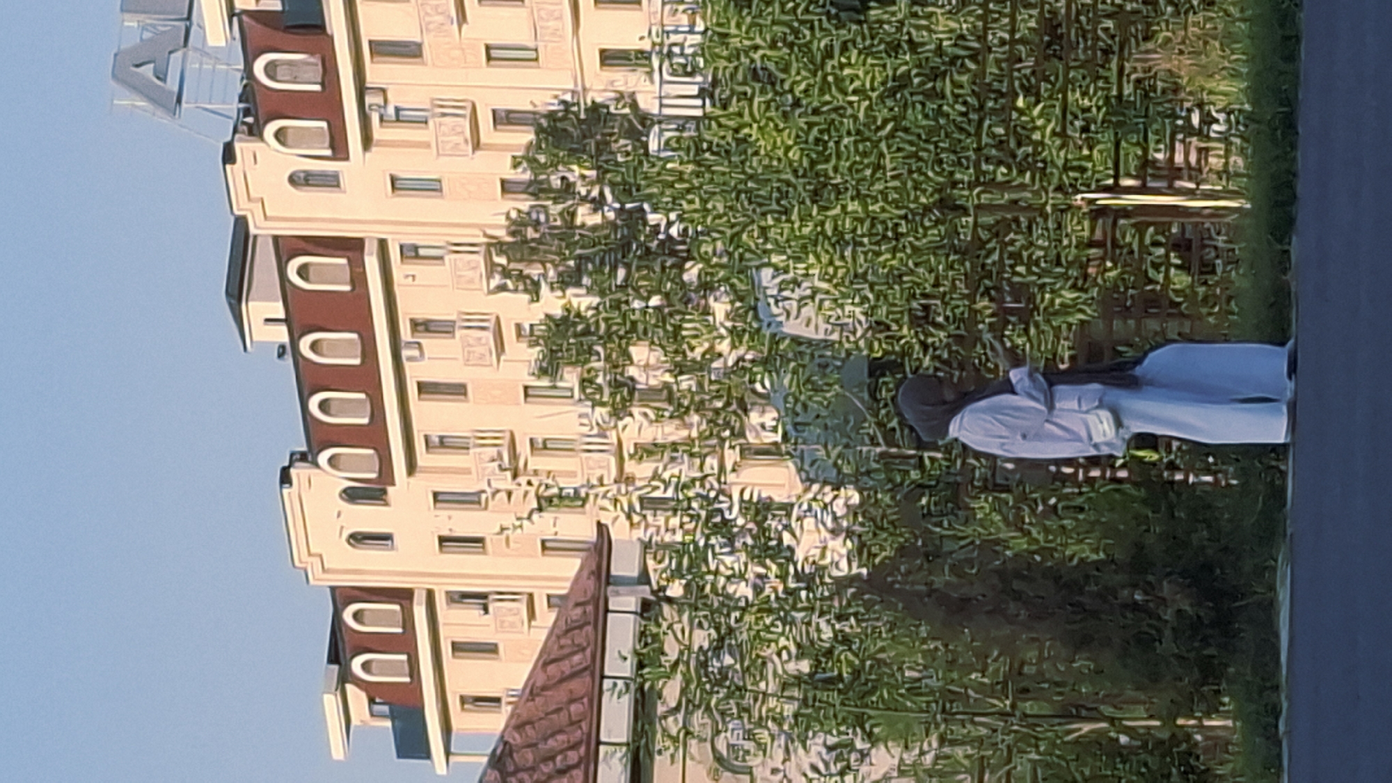 A full-body shot shows a person wearing a light-colored hijab, a white long-sleeved shirt, and loose white pants, standing in front of a hedge and a fence. The person is looking down at their phone, which they are holding in their right hand. A small white crossbody bag is hanging from their left shoulder. Behind the person, a beige building with arched windows and balconies is visible, with a large sign that appears to spell out "AVIA" in large silver letters against a clear blue sky. The building has a red-tiled roof. To the left of the building is a portion of a smaller building with a red-tiled roof. The scene appears to be outdoors, with the sun casting shadows from the trees. This location is identified as Fergana City, Uzbekistan.